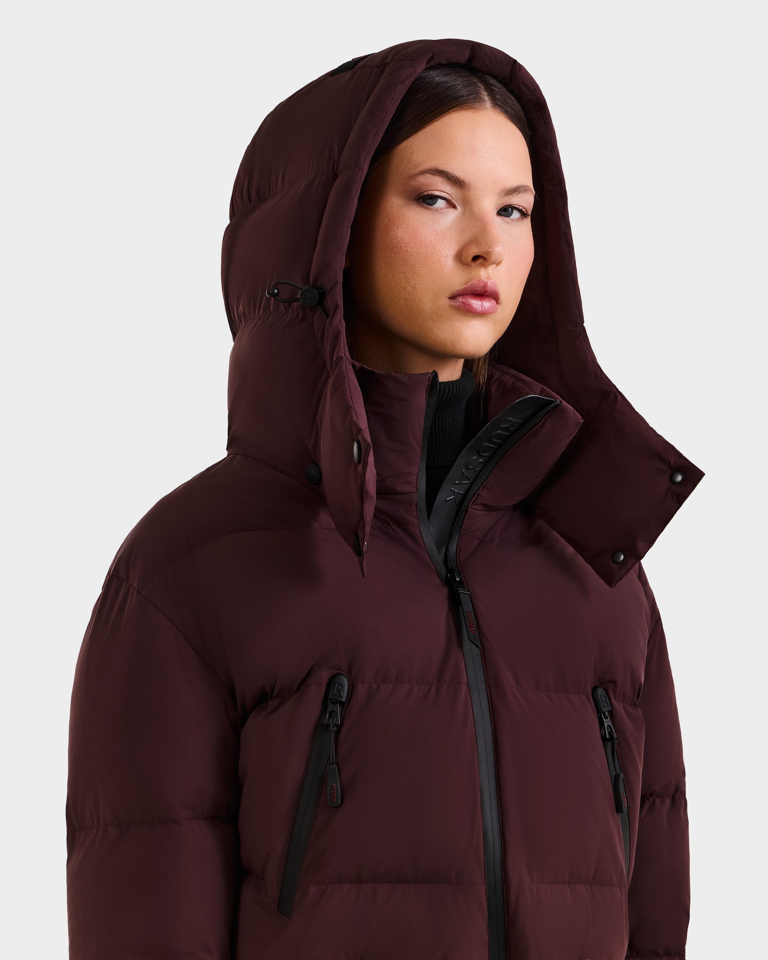 A woman wearing the Rudsak DINARA - 8125600 Bordeaux puffer jacket, with the hood up, gazes slightly to the side against a plain light background.