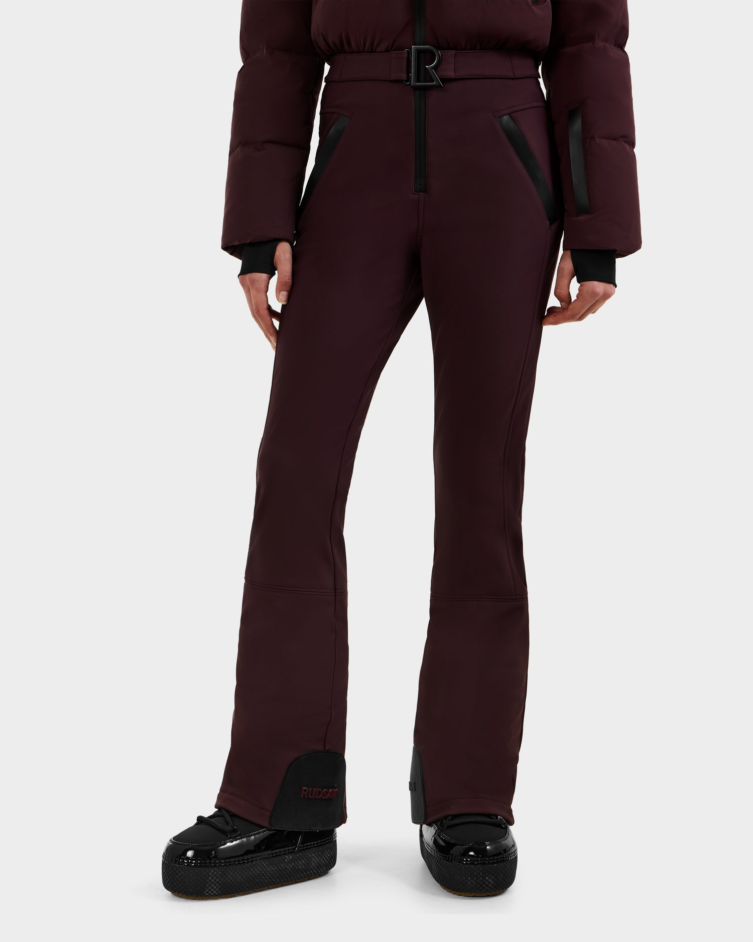 A person sports Rudsak’s DINARA - 8125600 Bordeaux ski pants with black zipper pockets and matching puffer jacket, showing off chic women’s ski suit style on the slopes; only the lower half is visible.