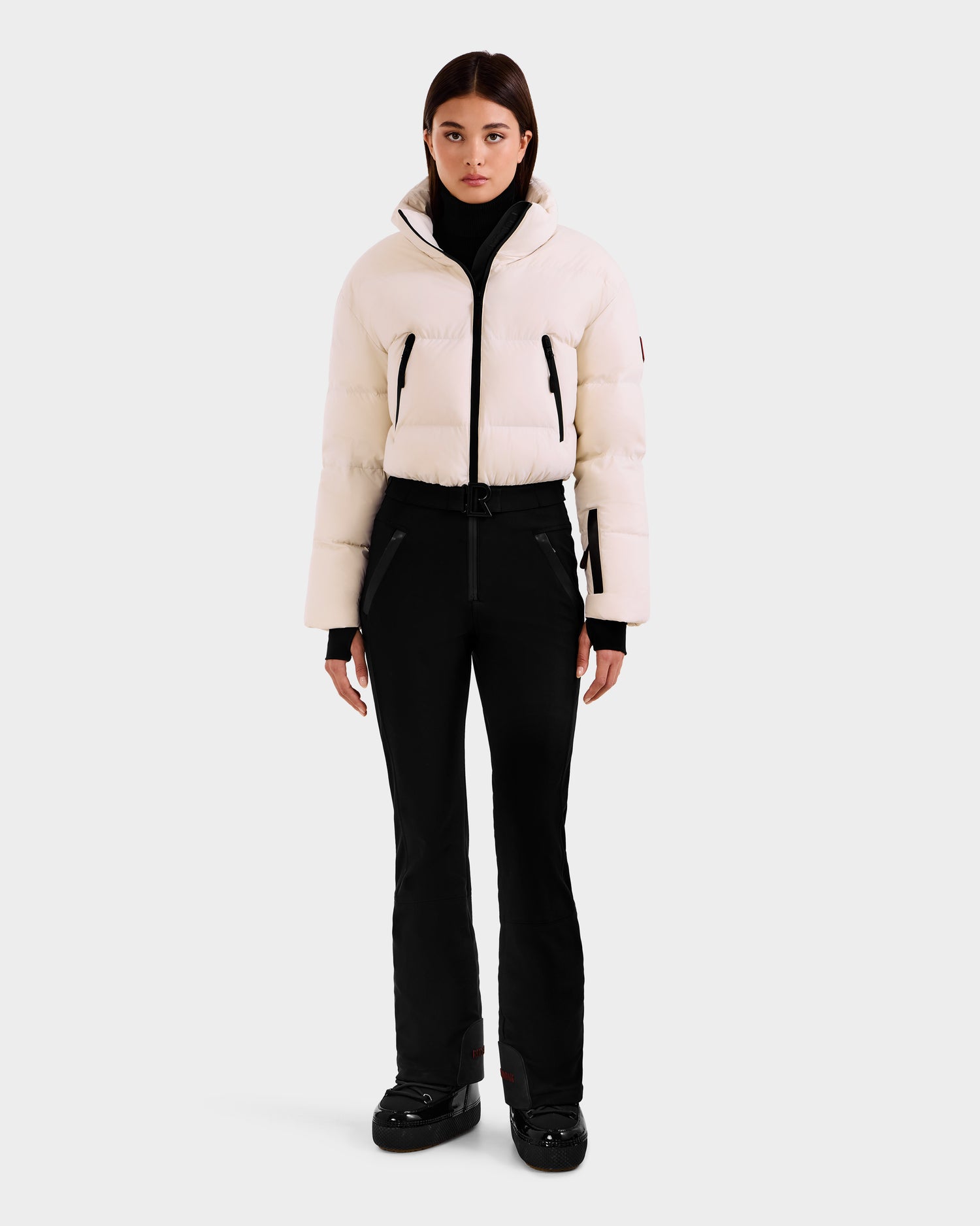 A woman stands facing forward in the Rudsak DINARA - 8125600 Vanilla women's ski suit with RECCO® rescue tech and black shoes, set against a plain light gray background.