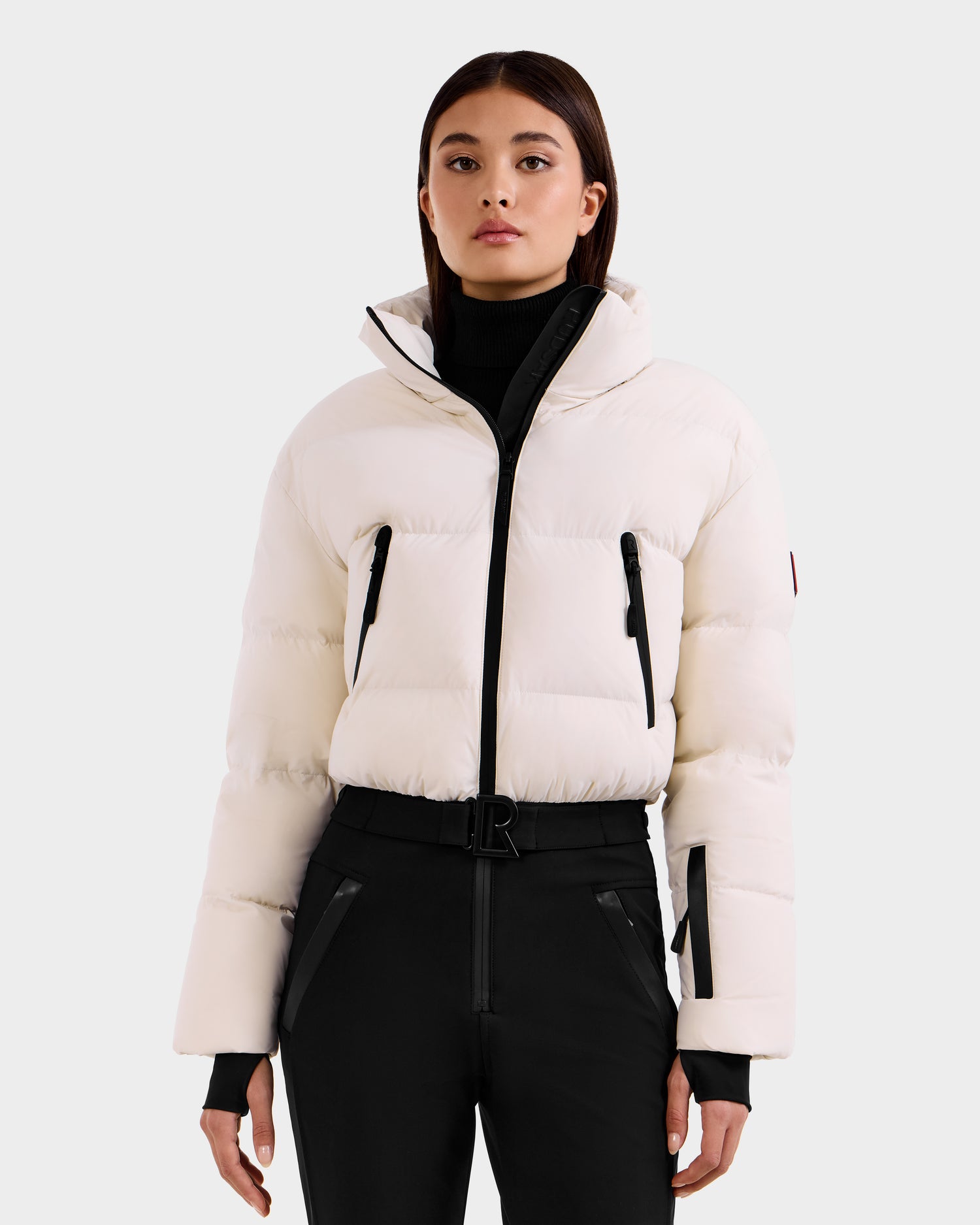 A woman wearing the Rudsak DINARA - 8125600 Vanilla white ski suit with black zippers, black high-waisted pants, and a black turtleneck stands facing forward against a plain light gray background.