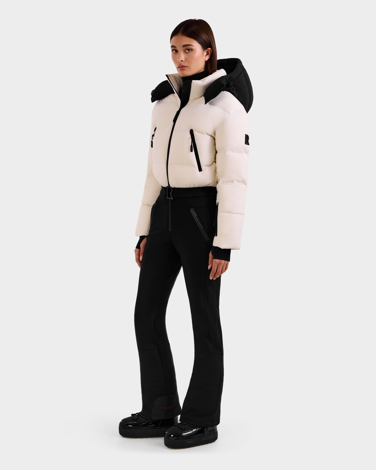 A woman faces forward wearing Rudsak’s DINARA - 8125600 Vanilla ski suit: a white high-collar puffer jacket with black accents, slim black pants, and black boots—an ideal winter or skiing outfit.