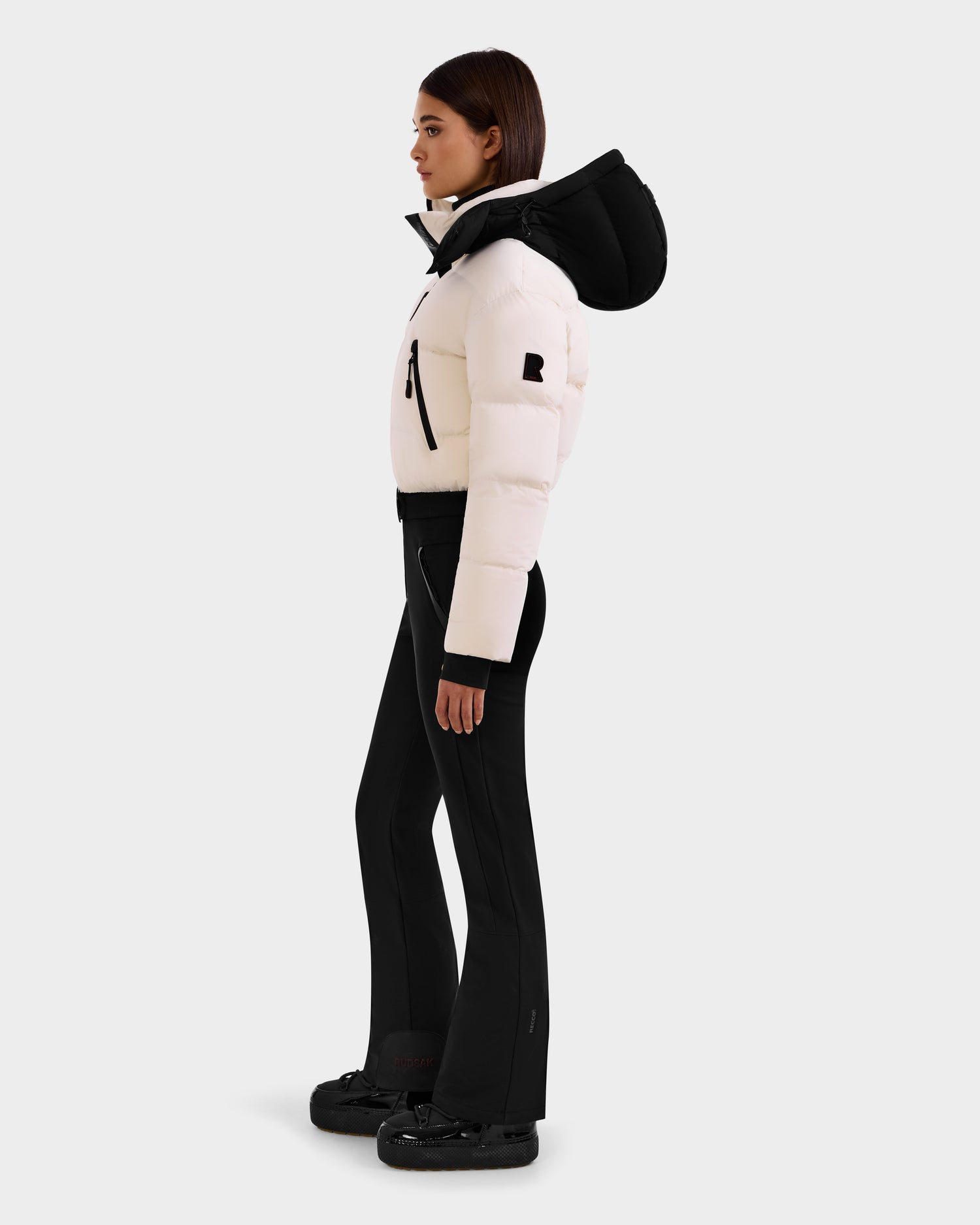 A woman stands in profile against a plain light background, wearing the Rudsak DINARA - 8125600 Vanilla puffer jacket with a black hood, paired with black flared pants and shoes for a modern, ski-inspired look.