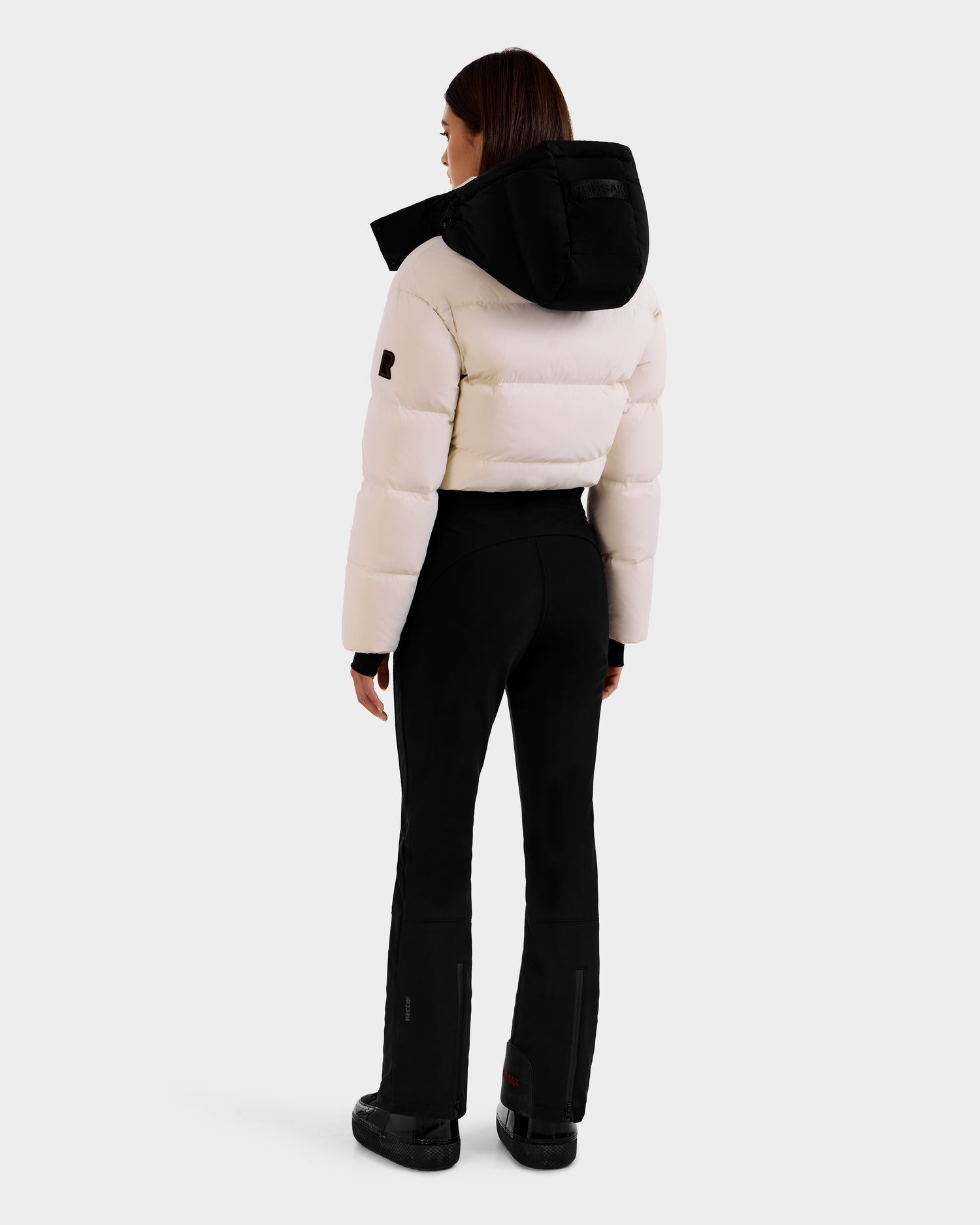 A woman faces away in the DINARA - 8125600 Vanilla white puffer jacket by Rudsak, featuring a black hood, black pants, and boots—her outfit equipped with RECCO® rescue technology for enhanced safety.
