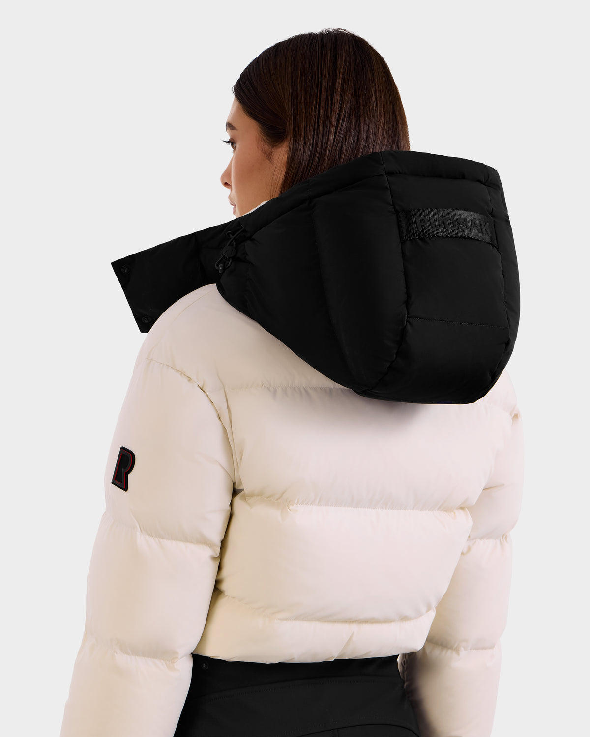 A person with straight brown hair is seen from behind wearing the Rudsak DINARA - 8125600 Vanilla women’s ski suit with a large black hood, showcasing its texture and details against a plain light background.