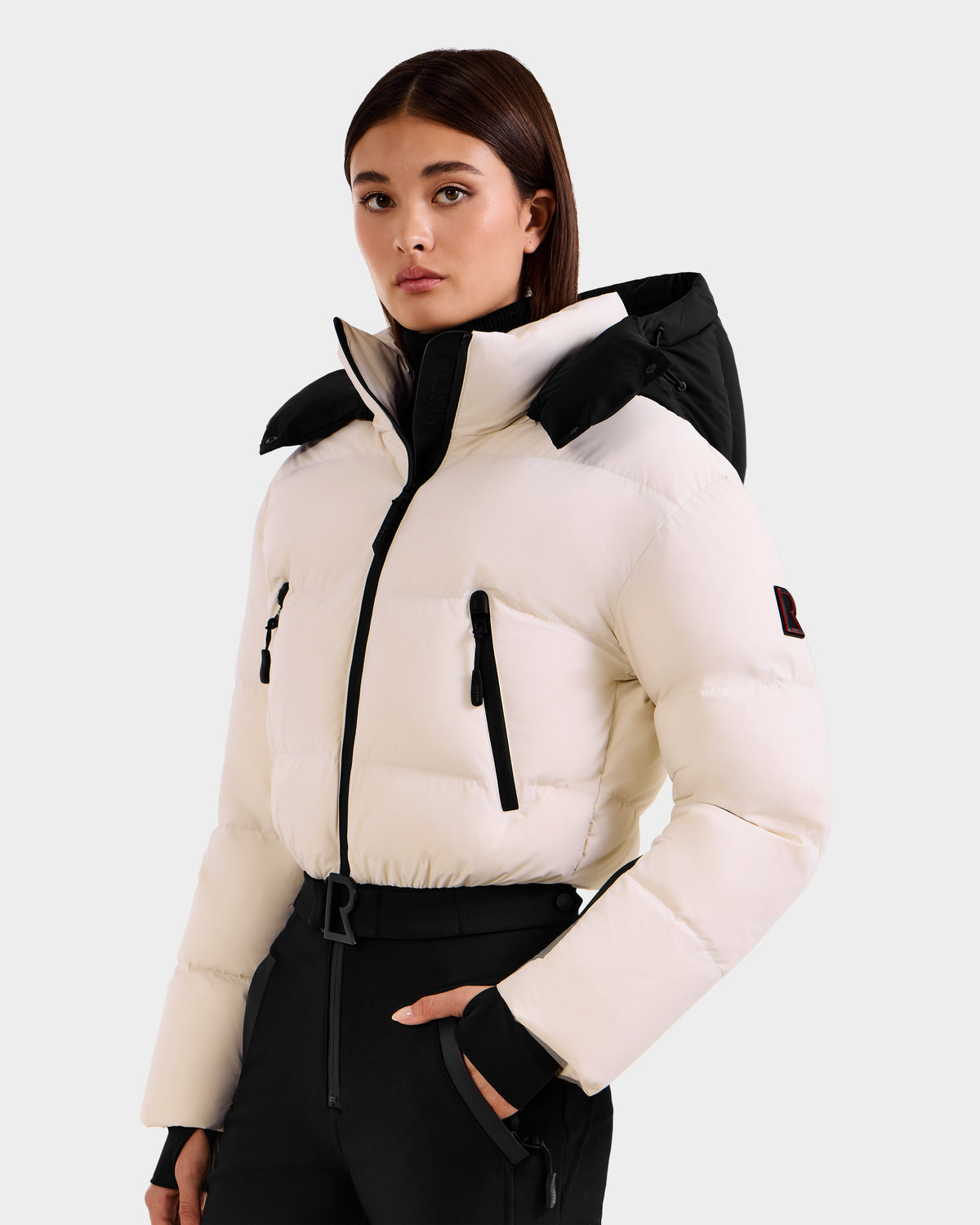 A woman models the Rudsak DINARA - 8125600 Vanilla ski suit, featuring a white puffer jacket with black zipper details and a black hood, standing confidently with one hand in her pocket against a plain light background.