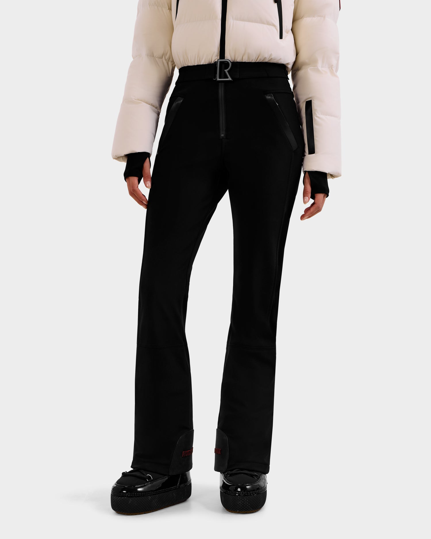 A person wearing the Rudsak DINARA - 8125600 Vanilla white puffer jacket, black fitted ski pants with RECCO® rescue tech and zippered pockets, and black snow boots stands against a plain white background.