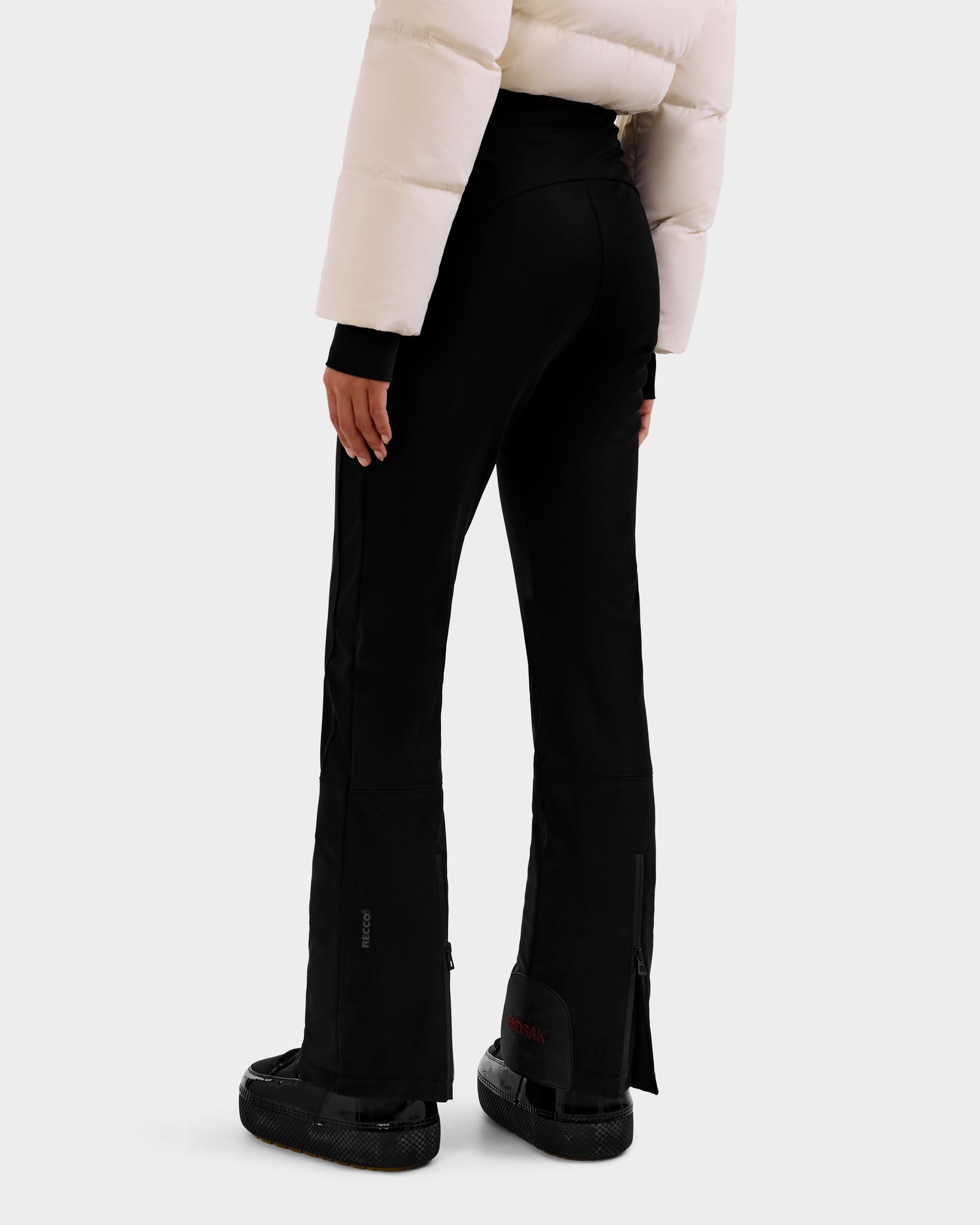 A person wearing Rudsak's DINARA - 8125600 Vanilla women's ski suit with RECCO® rescue technology, black flared pants, and black boots is shown from the back against a plain white background.