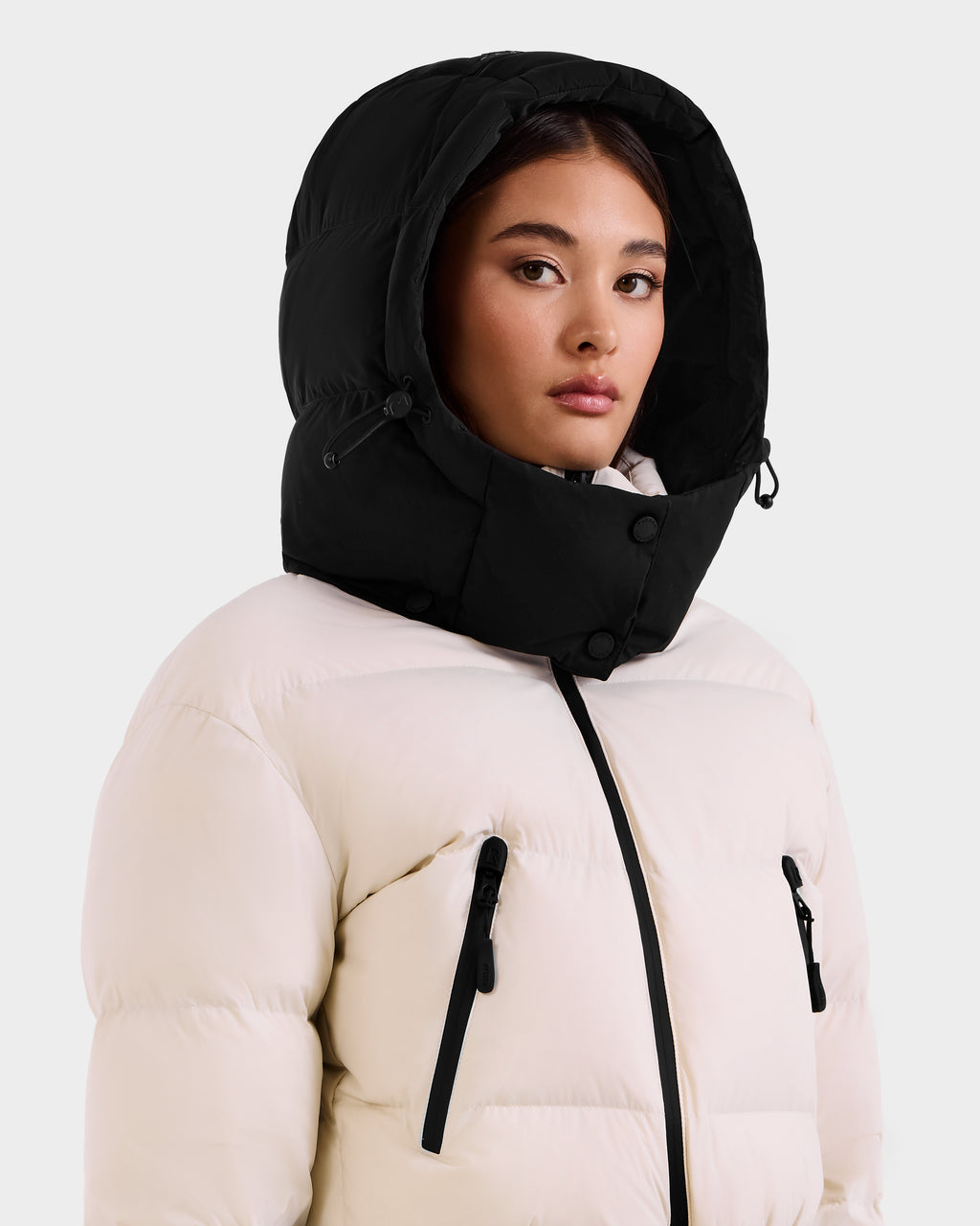 A woman wears the Rudsak DINARA - 8125600 Vanilla puffer jacket, featuring a large black hood, black zippers, and snap buttons. This ski suit–inspired design blends style and function against a plain light background.