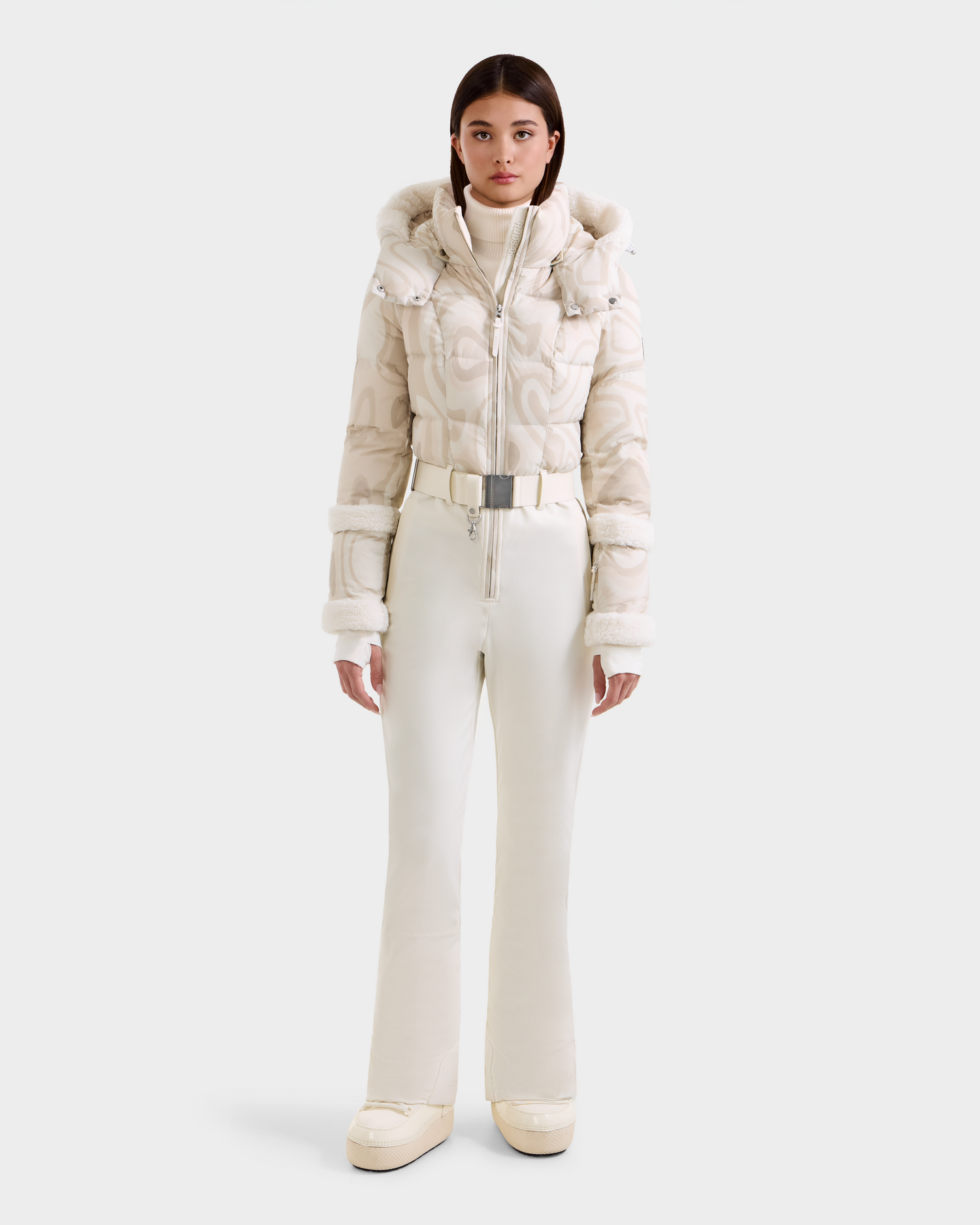 A woman models the Rudsak CARMEN P - 8125707 Vanilla Deja vu ski suit with a patterned puffer jacket, fur-trimmed hood, high-waisted belted pants, and platform boots. Available for pre-order; ships by November 30th.