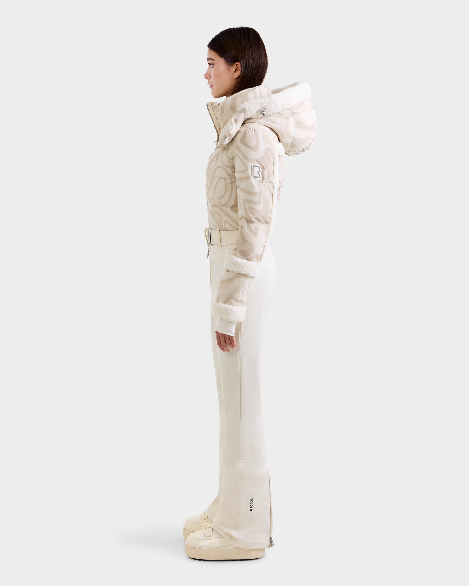 A woman stands in profile wearing the Rudsak CARMEN P - 8125707 Vanilla Deja vu ski suit with a patterned hooded jacket, belt, high-waisted pants, and matching snow boots. Pre-order now; expected to ship by November 30th.