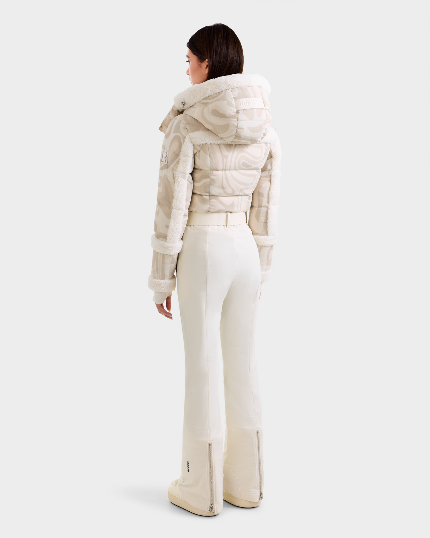 A woman faces away, wearing the Rudsak CARMEN P - 8125707 Vanilla Deja vu off-white quilted hooded jacket and high-waisted white ski pants. Fitted with subtle patterns. Pre-order now; ships by Nov 30. Plain light gray background.