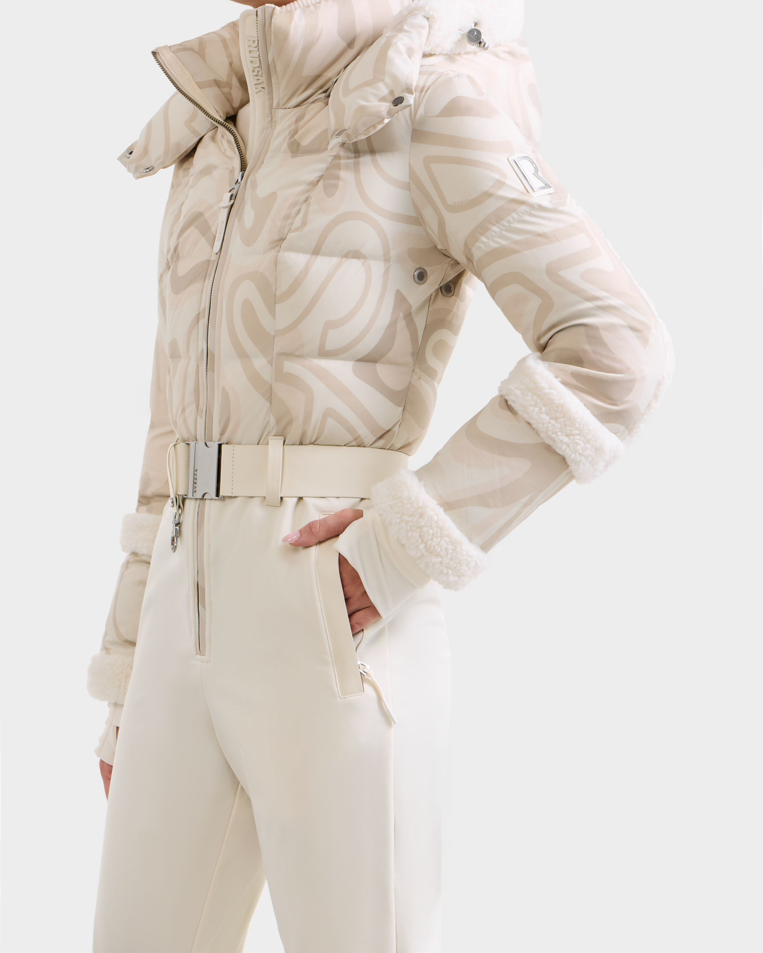 A person wears the Rudsak CARMEN P - 8125707 Vanilla Deja vu puffer jacket with a high collar and white fur trim, paired with cream high-waisted pants and matching belt—pre-order now, ships by November 30th.