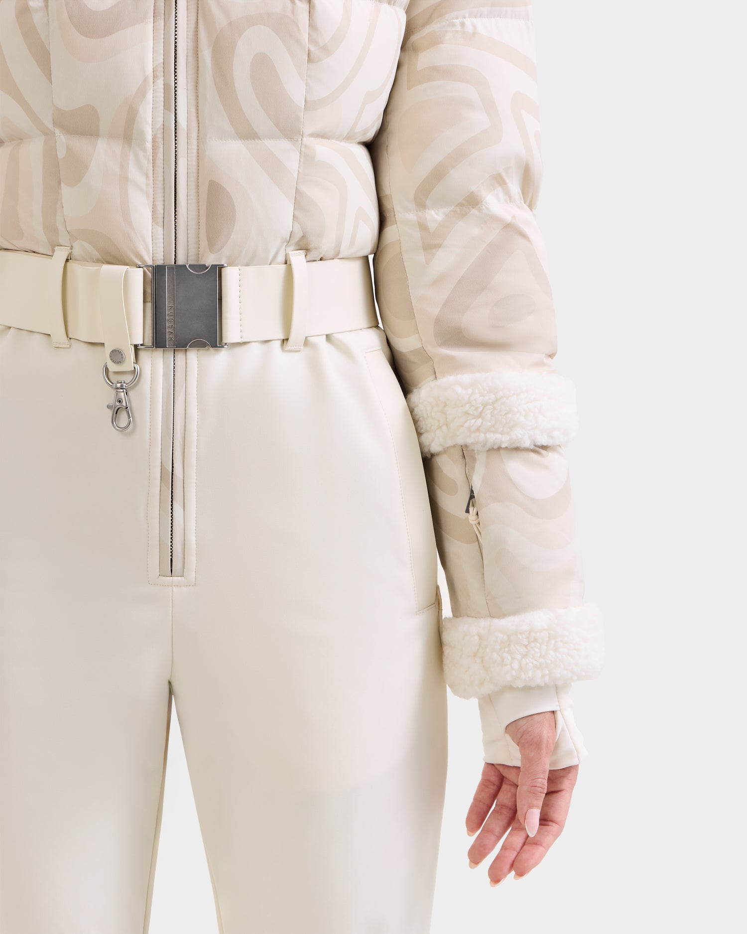 A person wears cream pants and a patterned light beige puffer jacket—CARMEN P - 8125707 Vanilla Deja vu by Rudsak—with white faux fur cuffs and a metallic-buckled belt. Available for pre-order, shipping by November 30th.