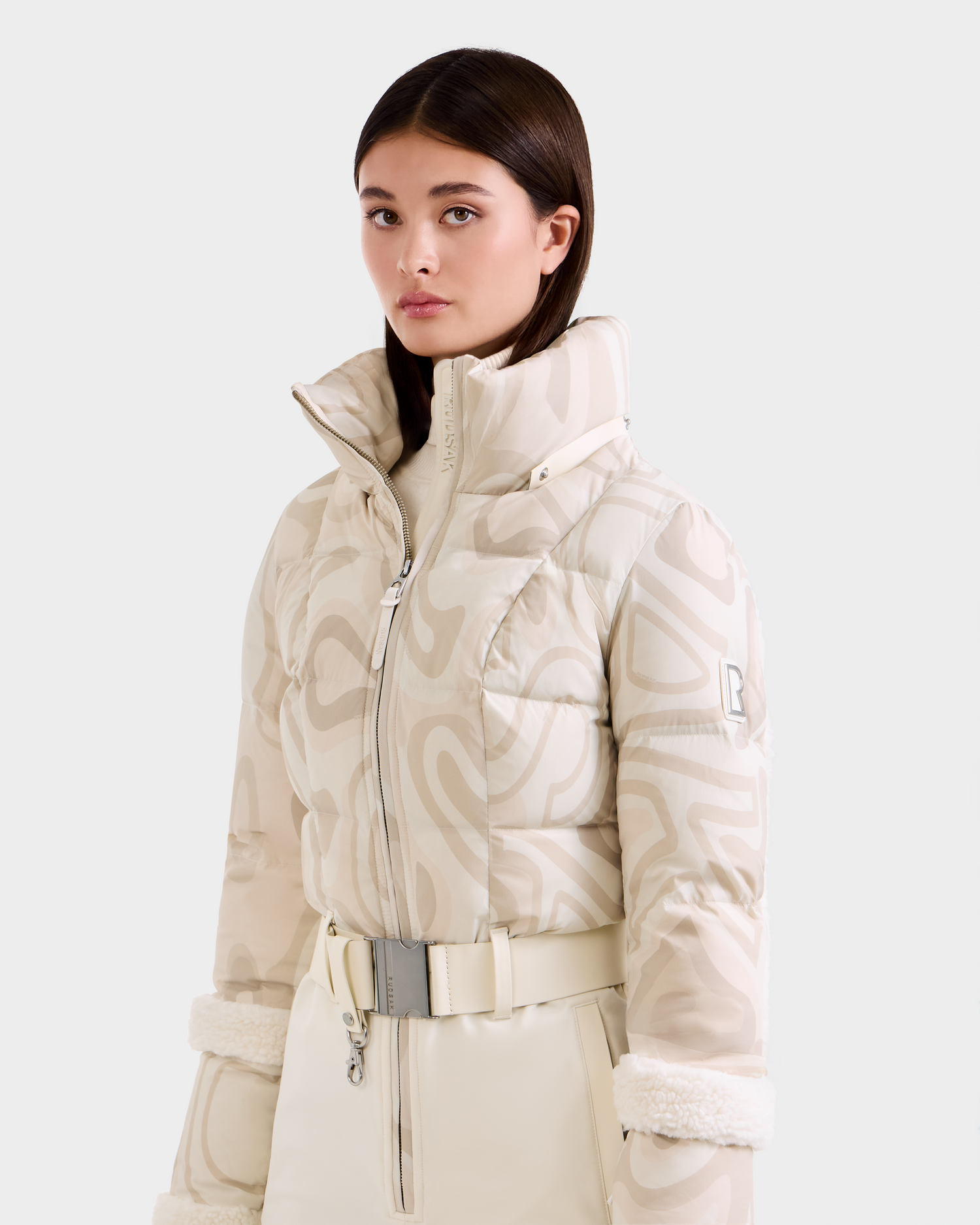 A woman with straight brown hair models the Rudsak CARMEN P - 8125707 Vanilla Deja vu: a high-collared, cream puffer jacket with matching belted pants, against a light gray background. This pre-order style ships November 30th.