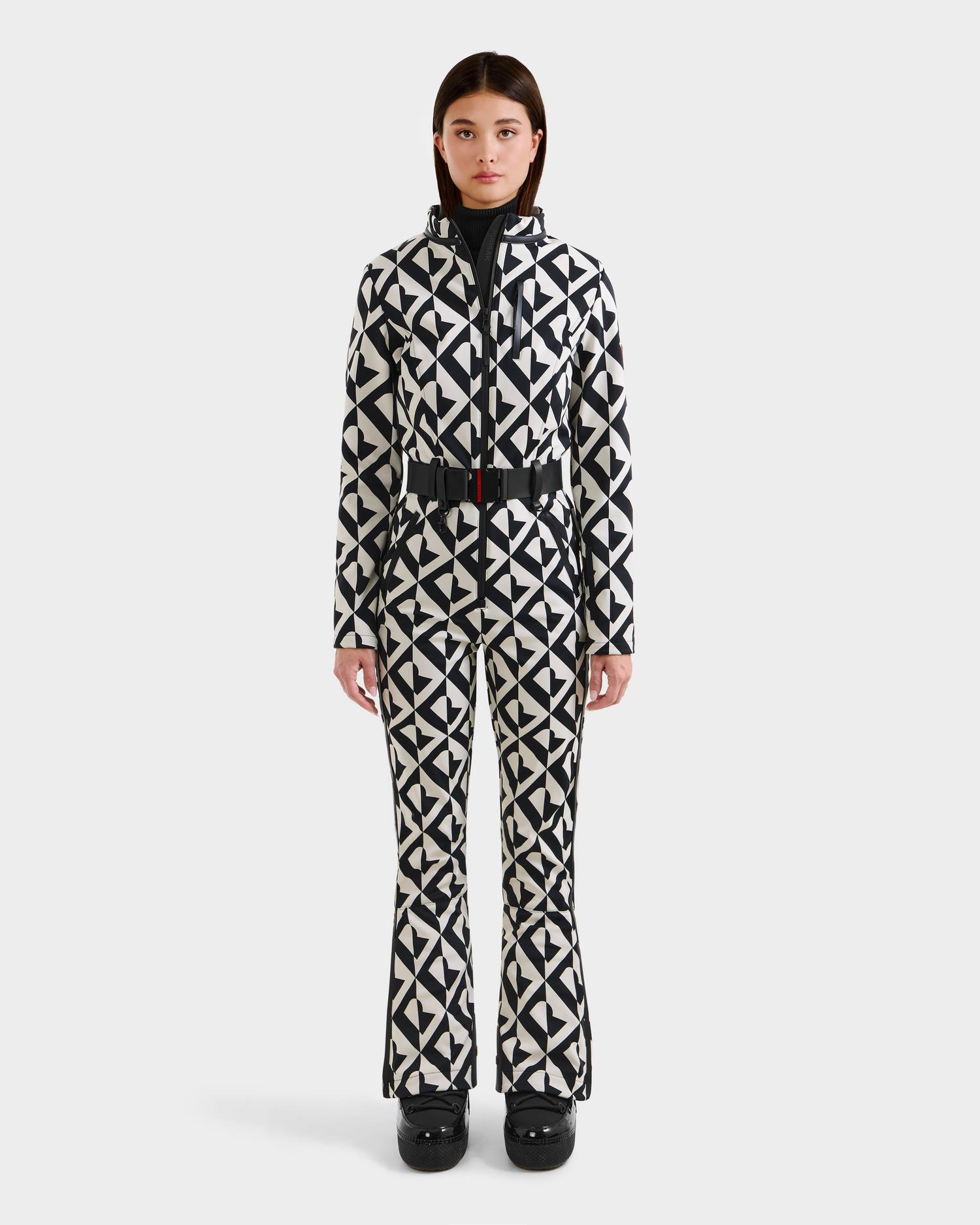 A person faces forward in the Rudsak JOSIE - 8124817 Vanilla Monogram women's one-piece ski suit with a belt, paired with black gloves and black ski boots, set against a plain white background.