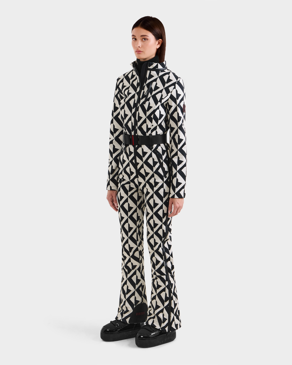 A person stands against a white background wearing the Rudsak JOSIE - 8124817 Vanilla Monogram women's ski suit with a black-and-white geometric pattern and black platform shoes.