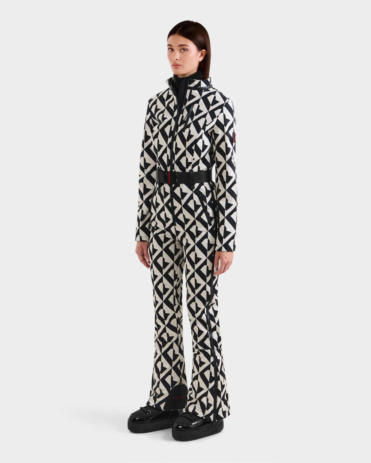 A person stands against a white background wearing the Rudsak JOSIE - 8124817 Vanilla Monogram women's ski suit with a black-and-white geometric pattern and black platform shoes.