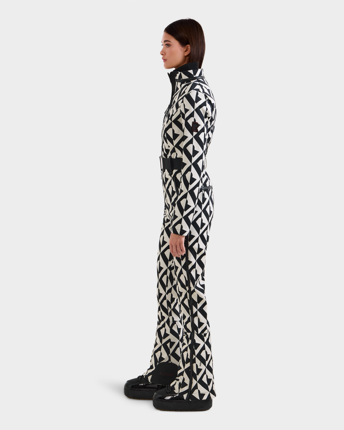 A woman in profile wears the Rudsak JOSIE - 8124817 Vanilla Monogram ski suit with a high collar, belted waist, and black boots, set against a plain white background.