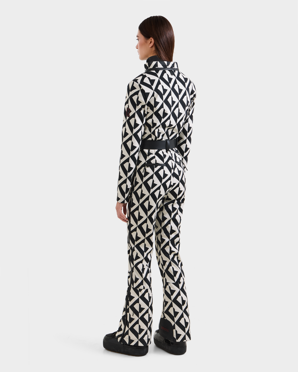 A woman faces away, wearing the Rudsak JOSIE - 8124817 Vanilla Monogram women's ski suit with a belt and black boots, set against a plain white background.