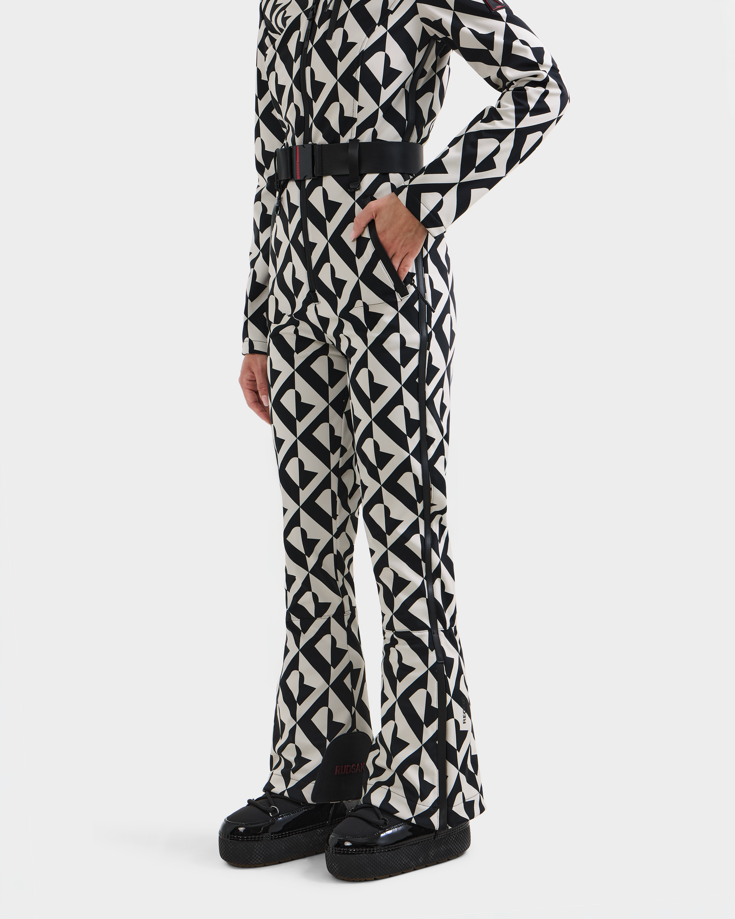 A person models the Rudsak JOSIE - 8124817 Vanilla Monogram women's ski suit with a belted waist and black platform shoes. The matching geometric outfit is shown from the neck down.
