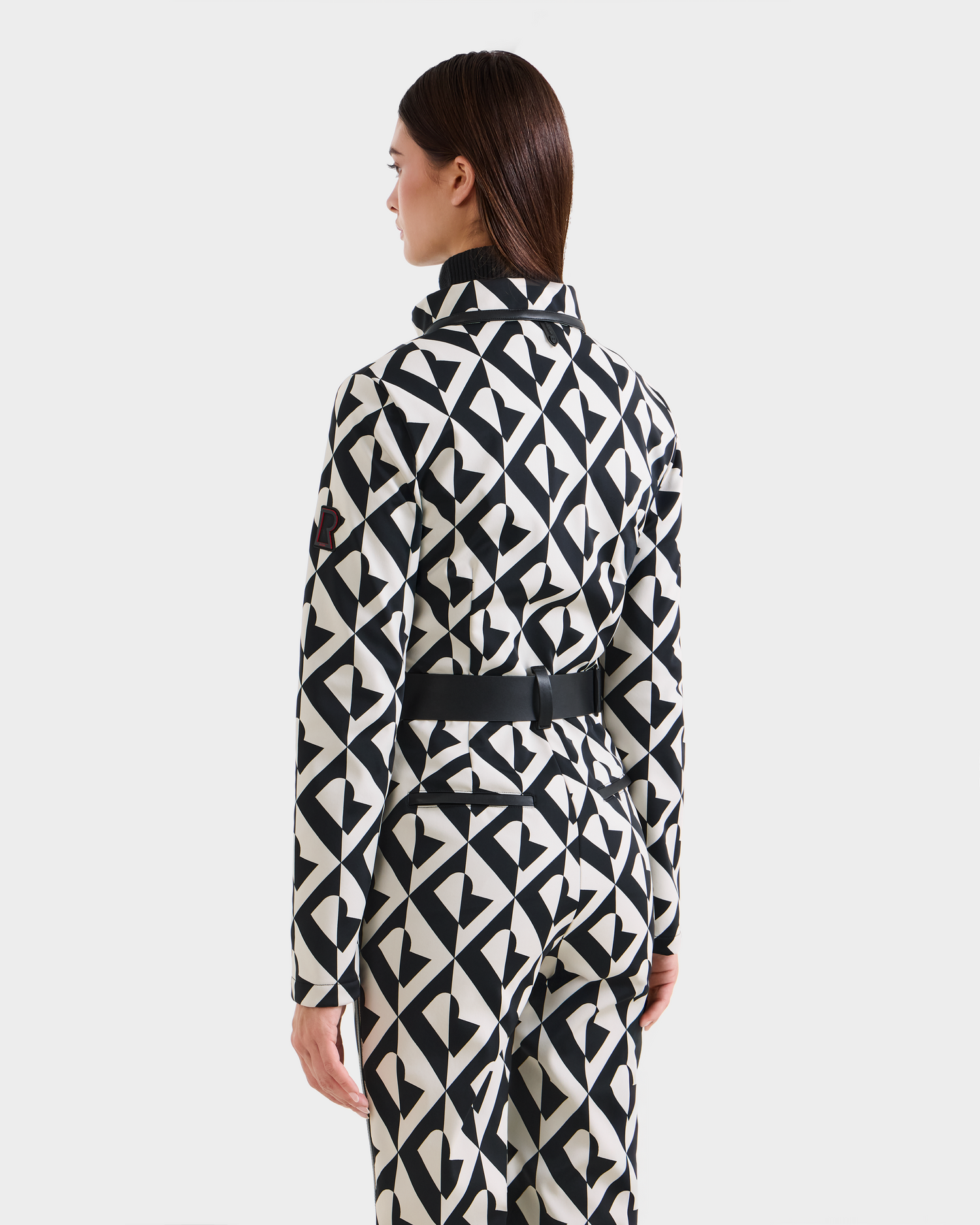 A woman faces away, wearing the Rudsak JOSIE - 8124817 Vanilla Monogram women's ski suit with a bold black and white geometric pattern, repeating triangle shapes, and a black belt at the waist. The background is plain and light.