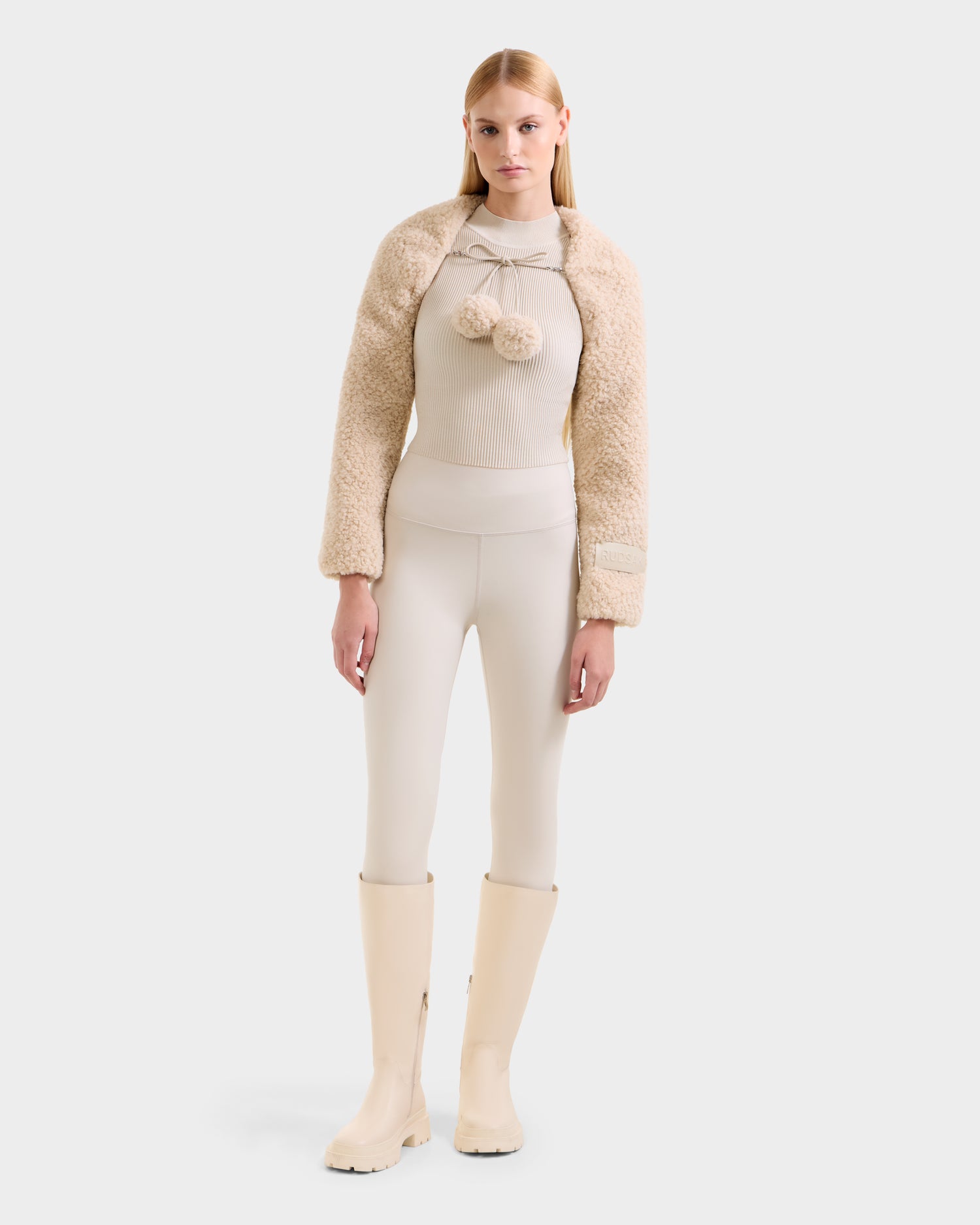 A woman faces forward, wearing the Rudsak VERITY - 8525666 Vanilla faux shearling bolero as a chic winter accessory over a beige top with pom-pom ties, matching fitted pants, and knee-high cream boots against a white background.