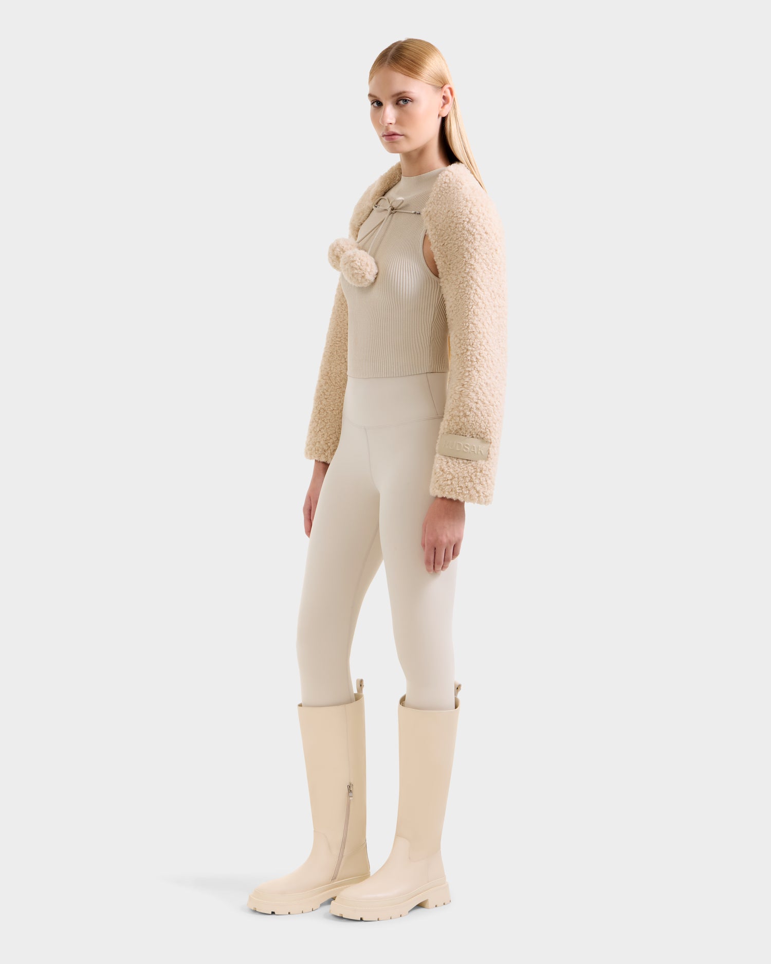 A woman with straight blonde hair wears the Rudsak VERITY - 8525666 Vanilla faux shearling bolero with pom-poms, styled with neutral separates and cream knee-high boots for an après-ski look against a light backdrop.