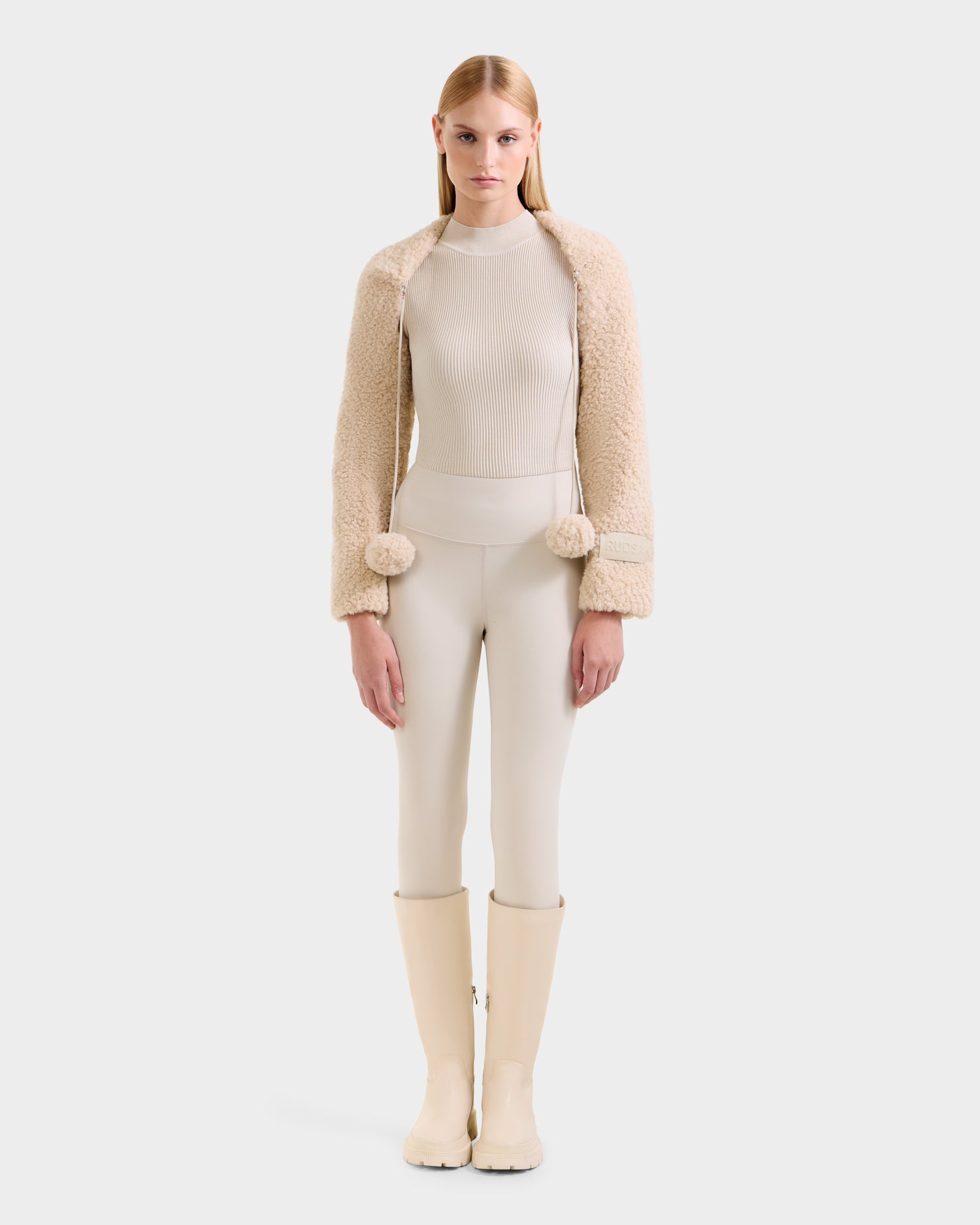 A woman faces forward, wearing the Rudsak VERITY - 8525666 Vanilla faux shearling bolero with pom-pom ties over a ribbed cream turtleneck, light fitted pants, and ivory knee-high boots against a plain white background.