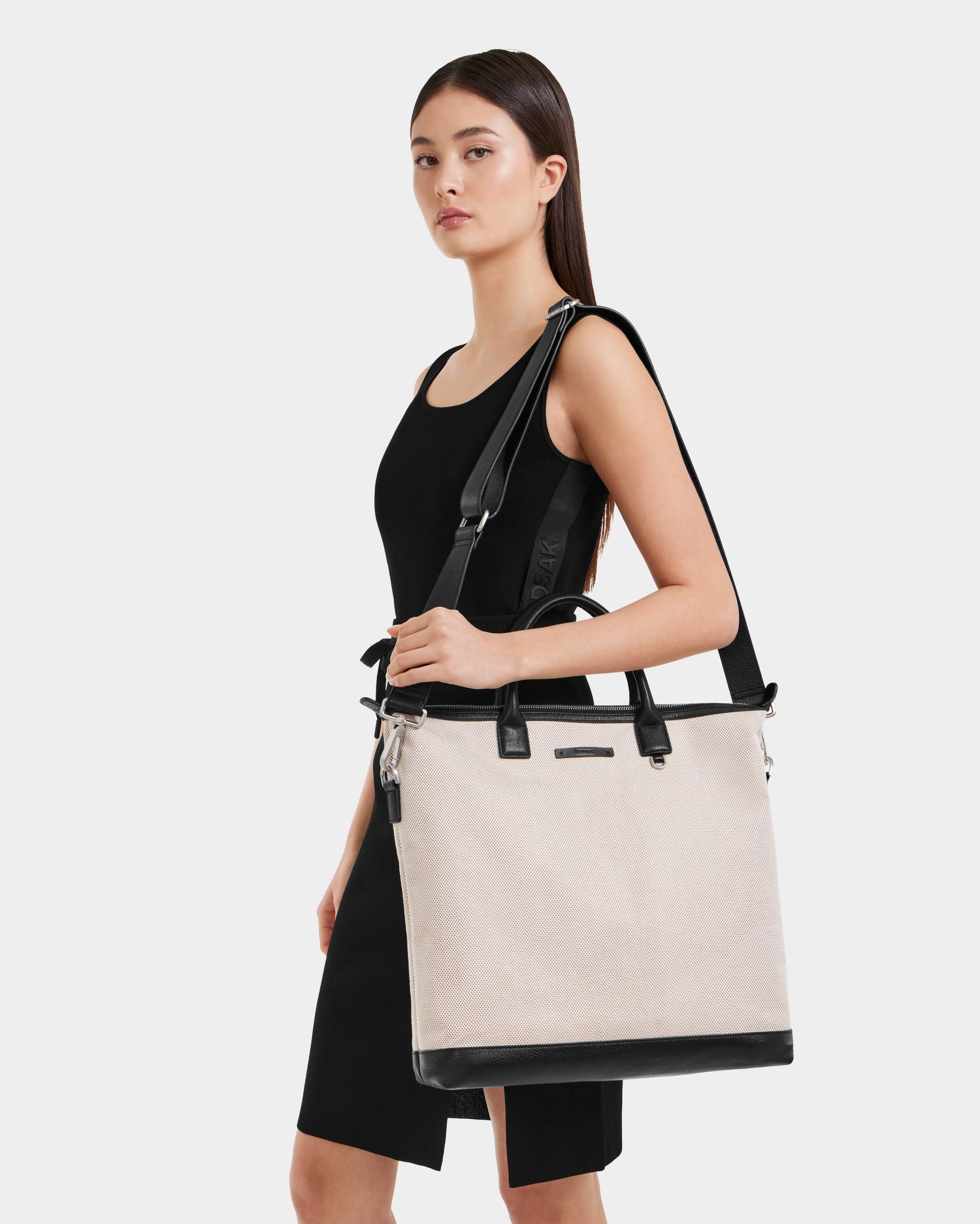 A woman in a sleeveless black dress holds the Rudsak DEXTER - 9324513 Vanilla, a large durable canvas tote with black handles, accents, and a shoulder strap, standing against a plain white background.