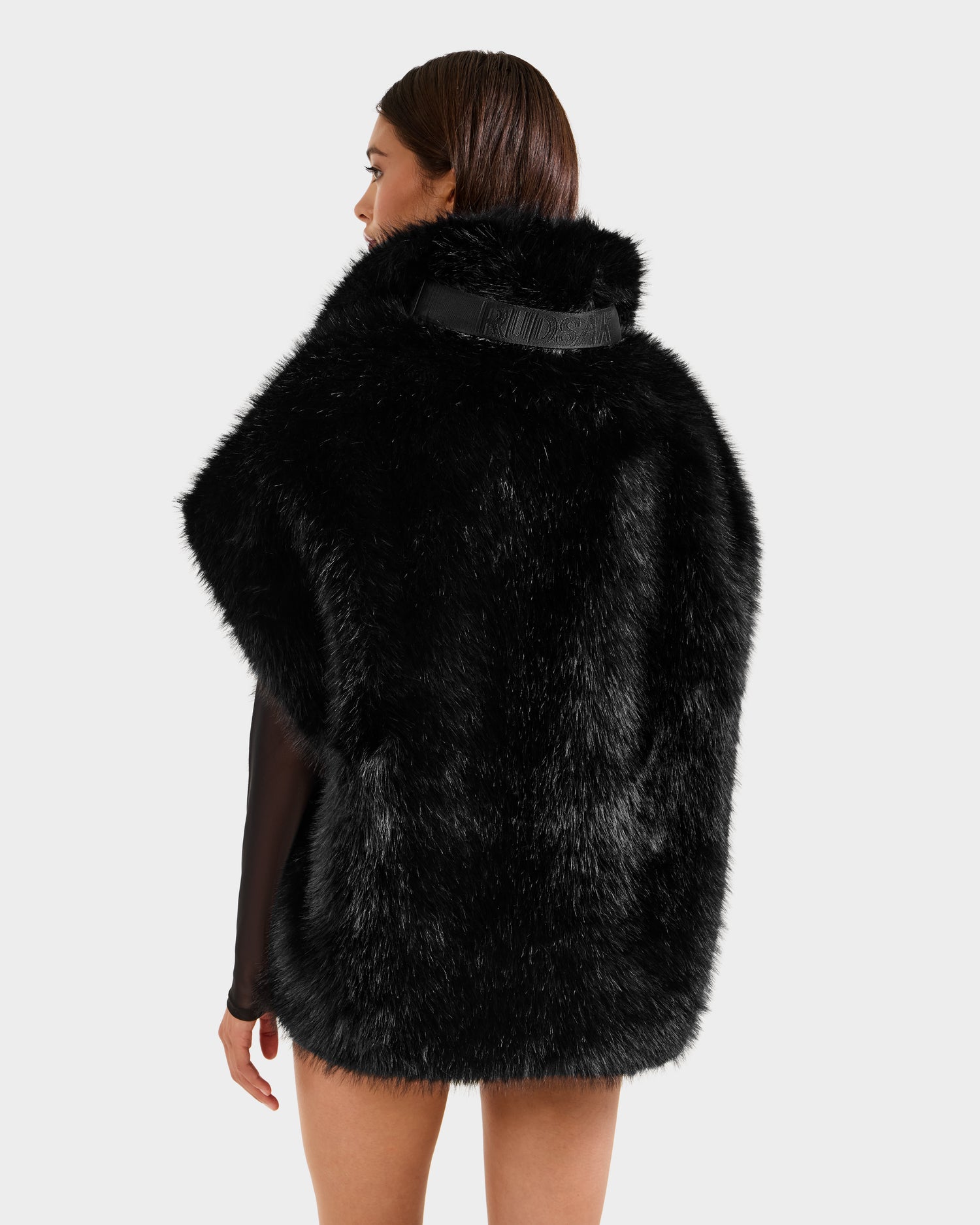 A woman with straight brown hair wears the Rudsak DELFINA - 8525662 Black short faux fur cape, its oversized silhouette adding drama to winter style, seen from behind against a plain light gray background.
