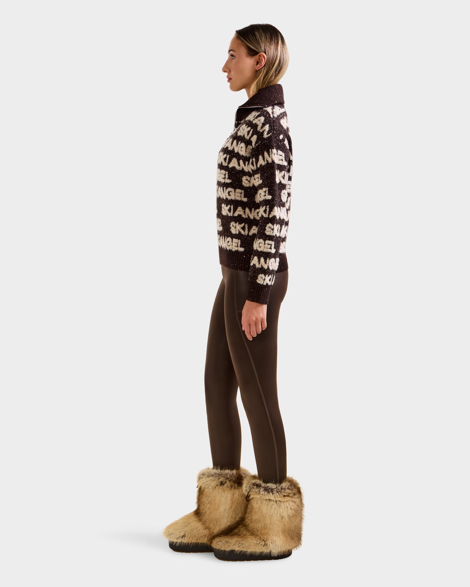 A woman stands in profile against a plain white background, wearing the Rudsak LYDIA - 8525687 Espresso sweater, paired with brown leggings and tan furry boots.