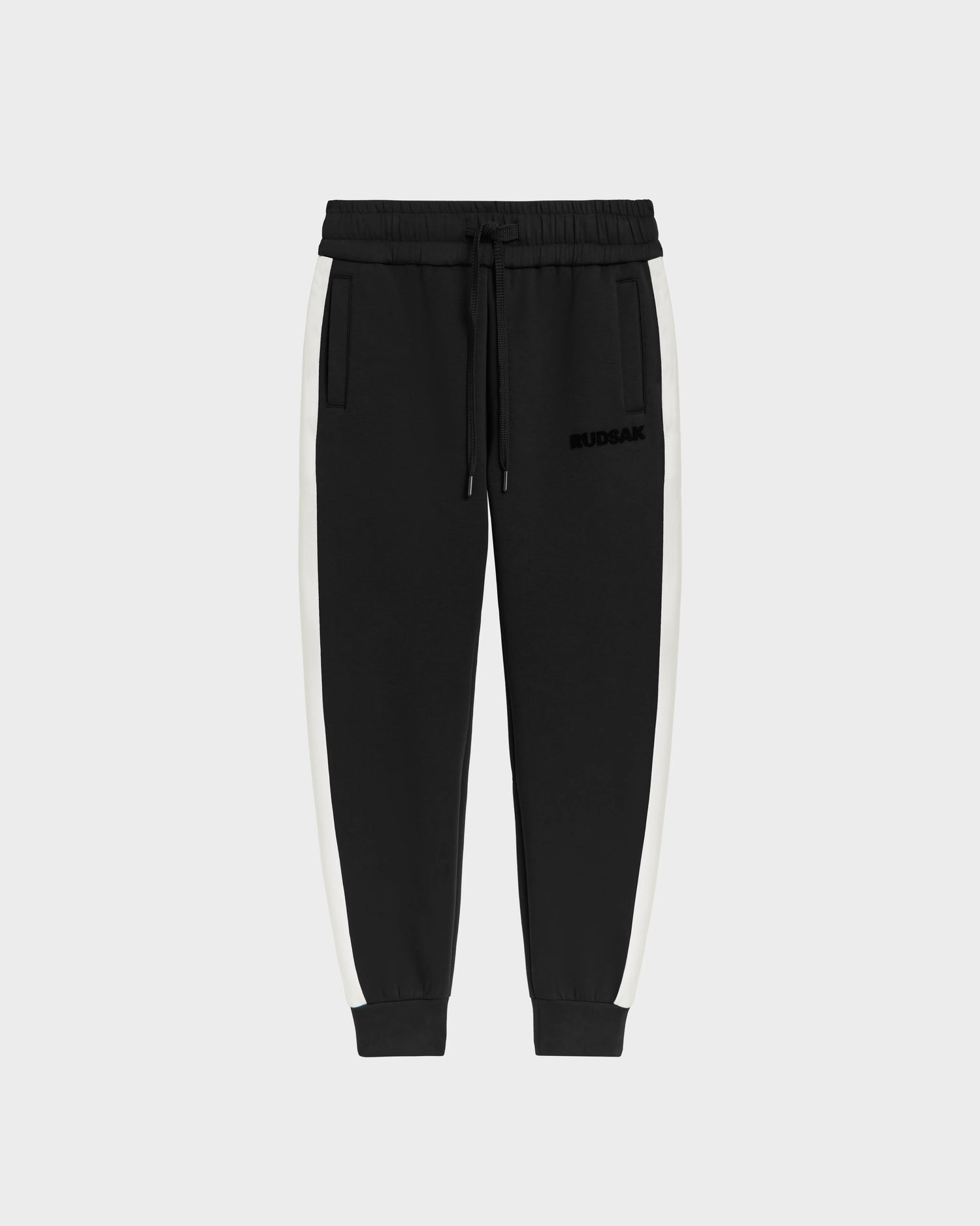 Rudsak RYLAN - 6525009 Black cotton-blend joggers feature white side stripes, an elastic drawstring waistband, side pockets, ribbed cuffs, and a subtle left thigh logo, shown on a plain light background.