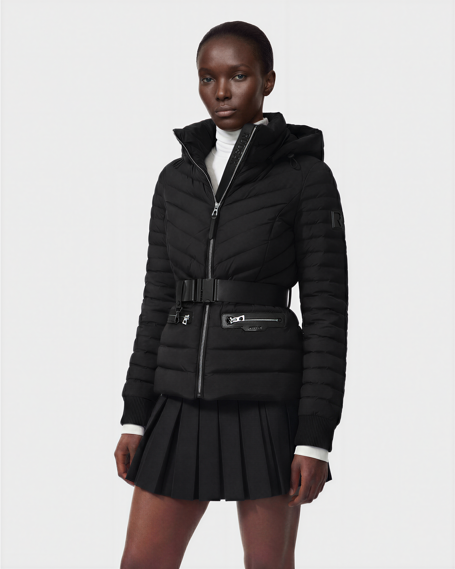 A person wears the Rudsak SCARLETT M - 8125565 Black water-resistant quilted jacket with a belt, zip pockets, and hood, styled over a white turtleneck and black pleated skirt, standing against a plain light background.