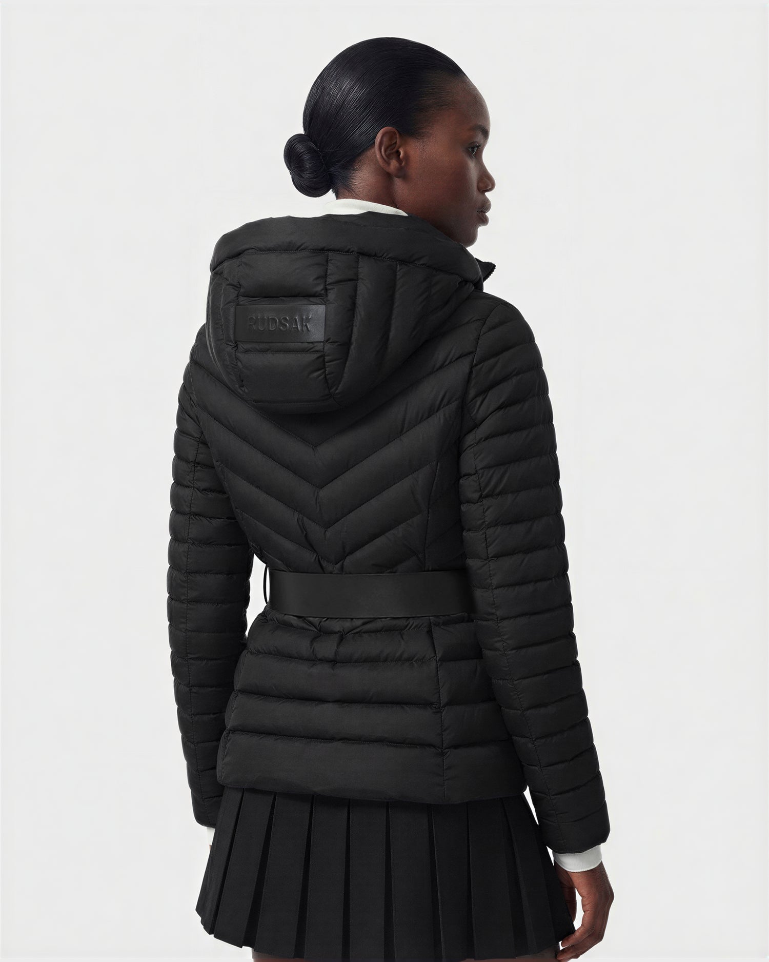 A woman with neatly styled hair wears Rudsak’s SCARLETT M - 8125565 Black, a women’s lightweight water-resistant down jacket with a belt, paired with a pleated black skirt, standing against a plain light background.