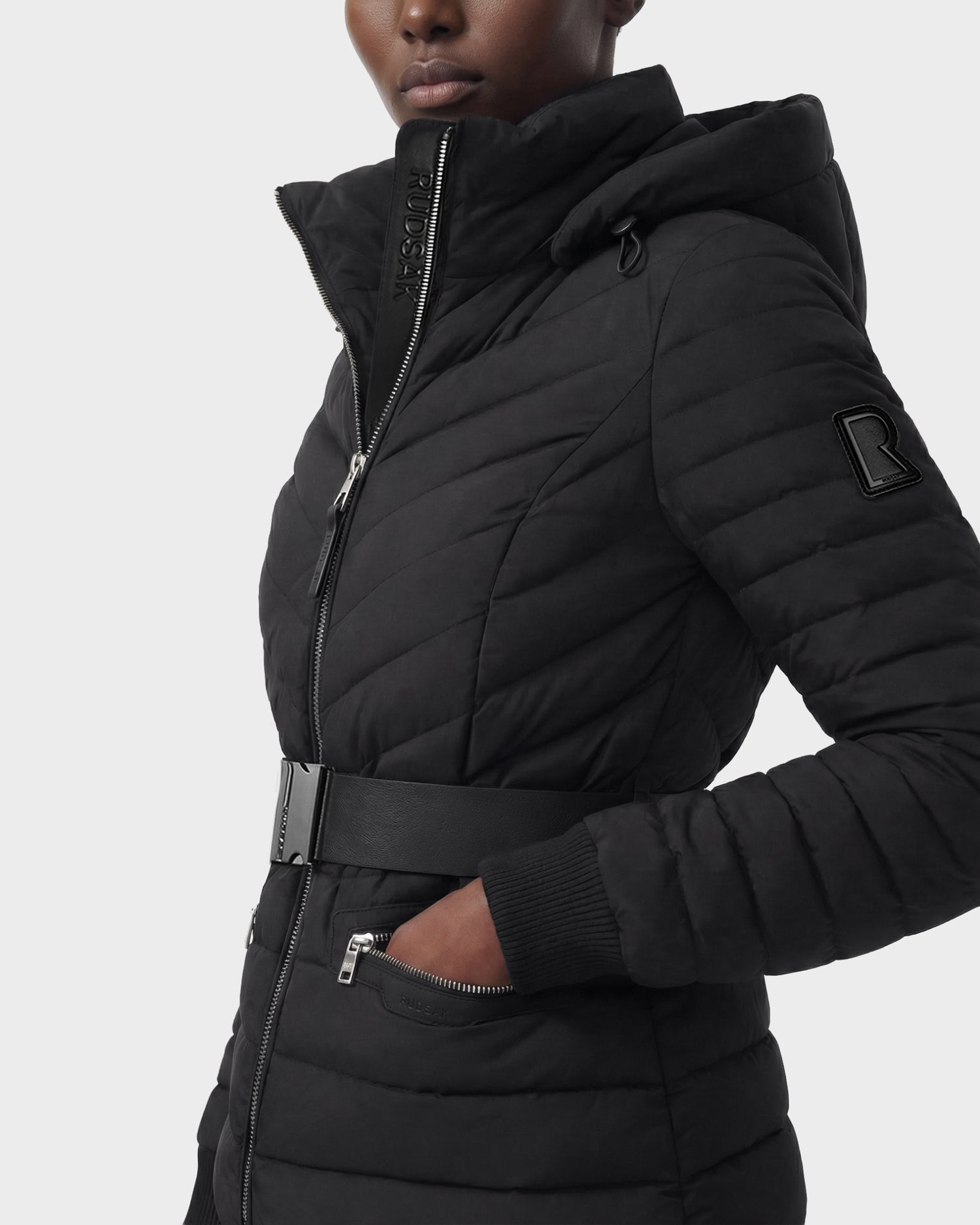A person wearing the Rudsak SCARLETT M - 8125565 Black, a black women's quilted lightweight down jacket with a belted waist, high collar, and zippered pocket, shown from shoulders to mid-thigh against a plain background.