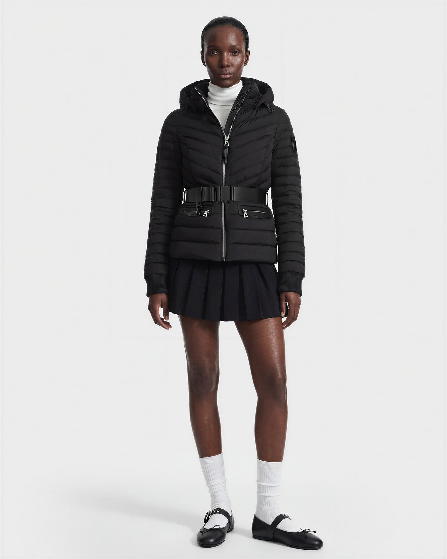 A person wears the Rudsak SCARLETT M - 8125565 Black women's lightweight down jacket with a black belt, white turtleneck, black pleated skirt, white socks, and black shoes against a plain white background.