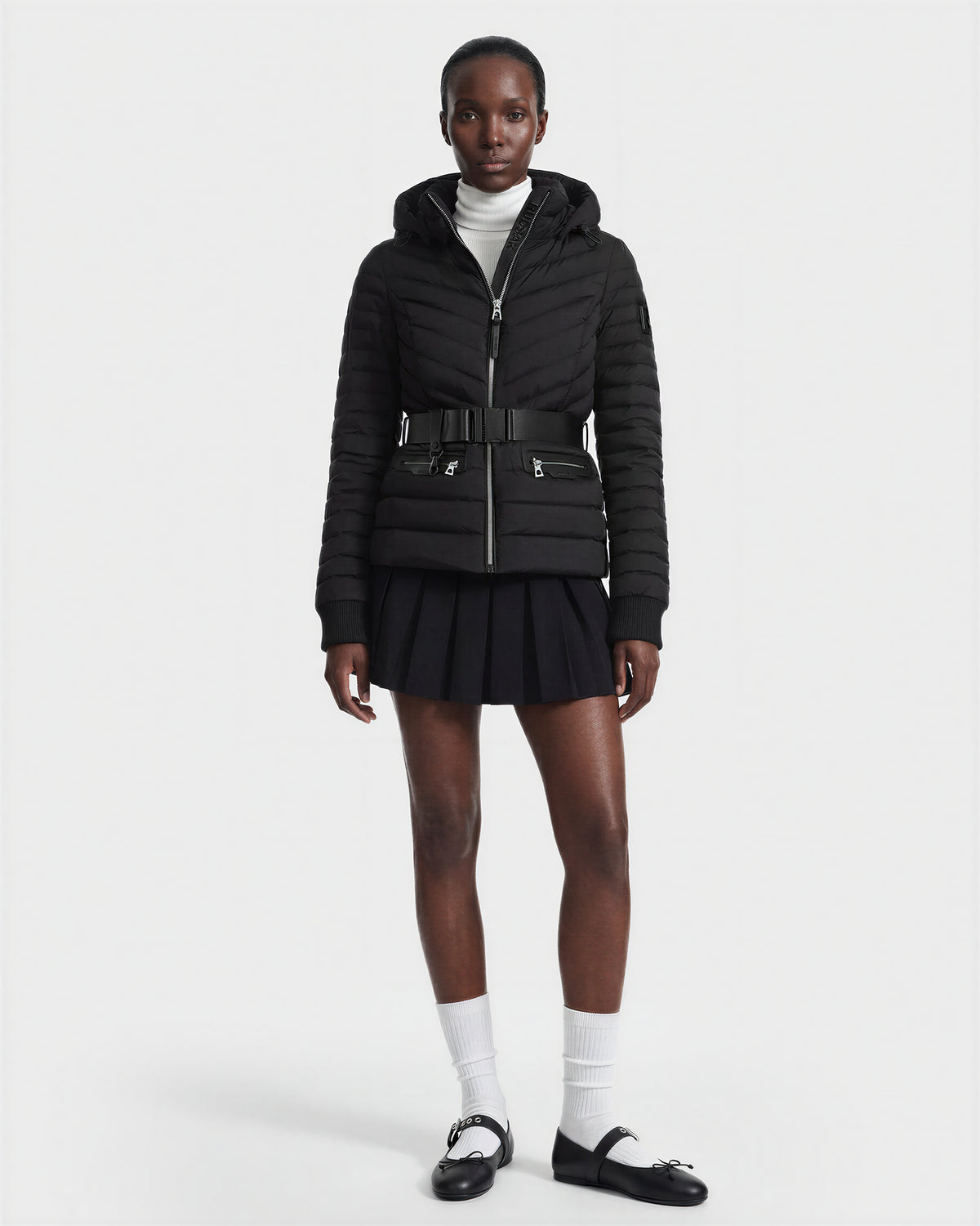 A person wears the Rudsak SCARLETT M - 8125565 Black women's lightweight down jacket with a black belt, white turtleneck, black pleated skirt, white socks, and black shoes against a plain white background.
