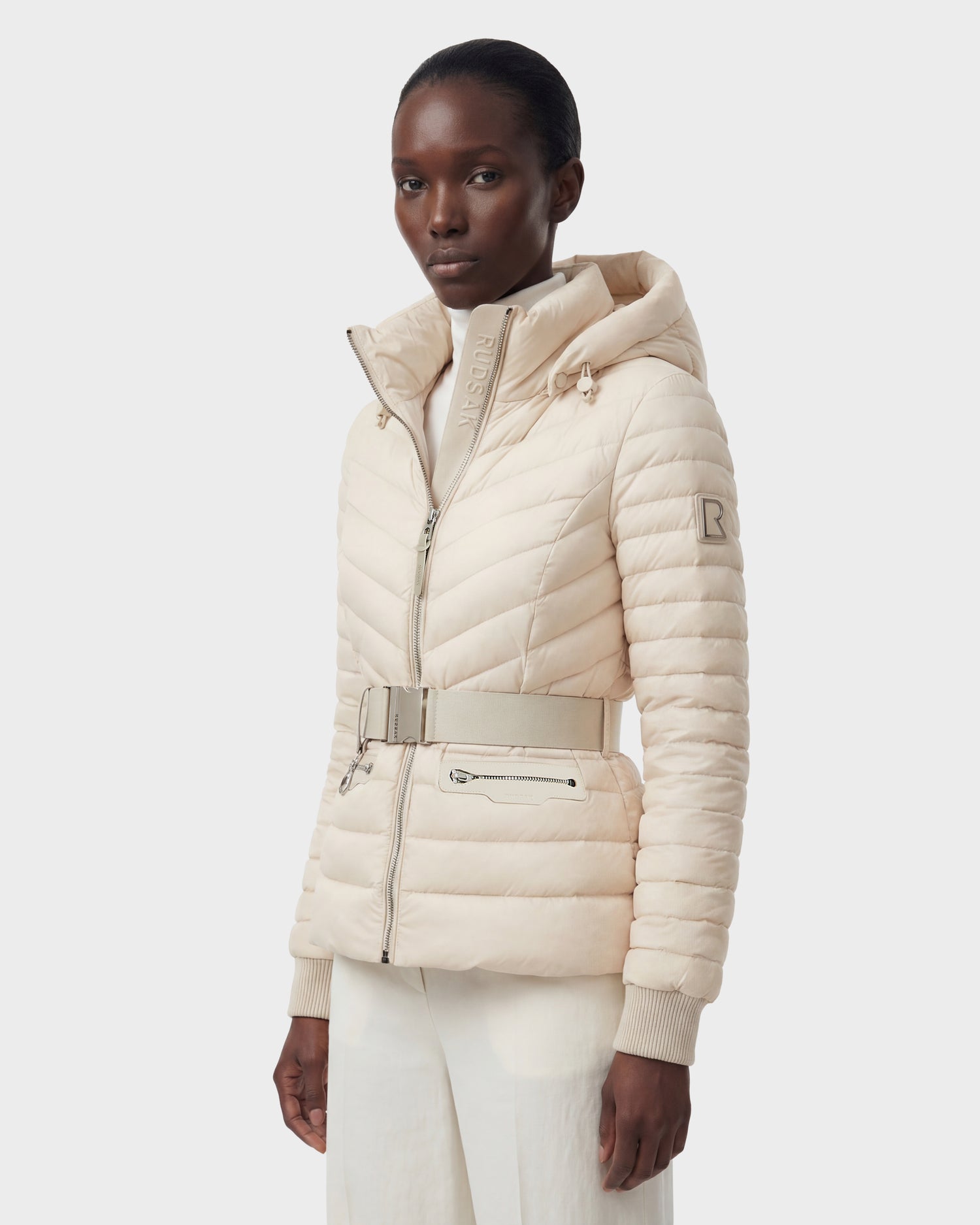 A person wears the Rudsak SCARLETT - 8125004 Vanilla, a light beige women’s quilted down jacket with a vegan leather belt and zip closure, paired with white pants, standing against a plain light background.