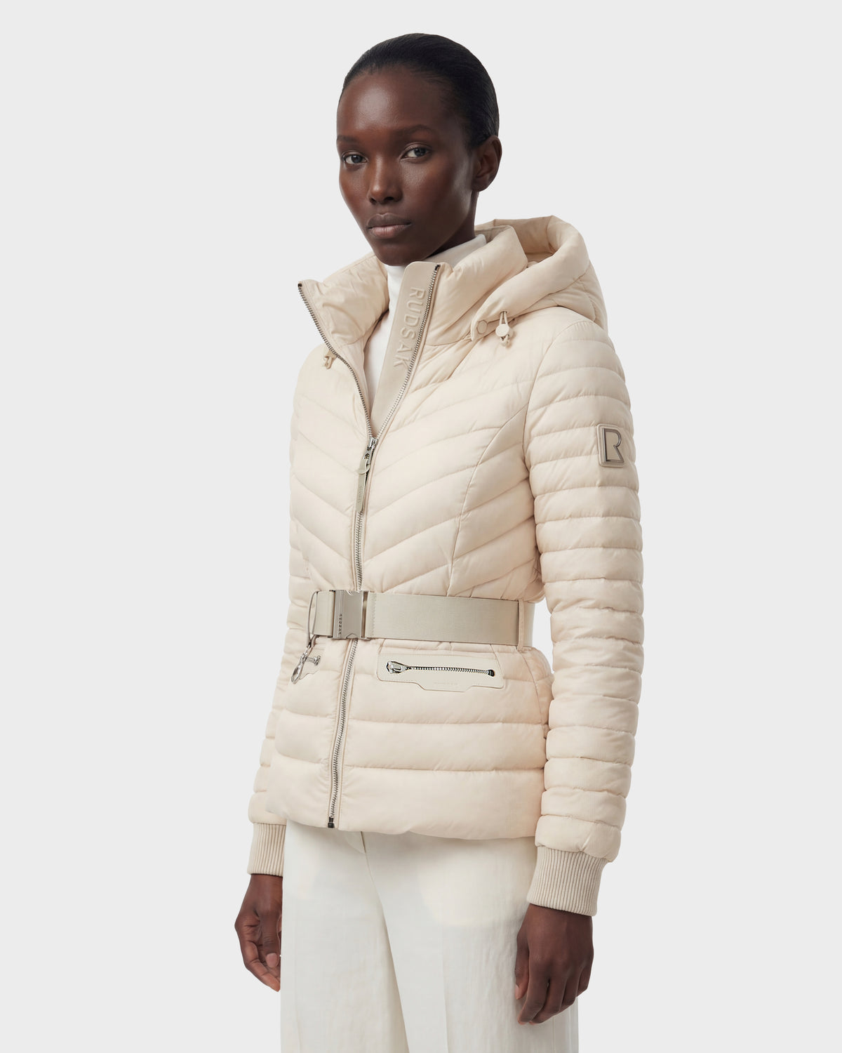 A person wears the Rudsak SCARLETT - 8125004 Vanilla, a light beige women’s quilted down jacket with a vegan leather belt and zip closure, paired with white pants, standing against a plain light background.