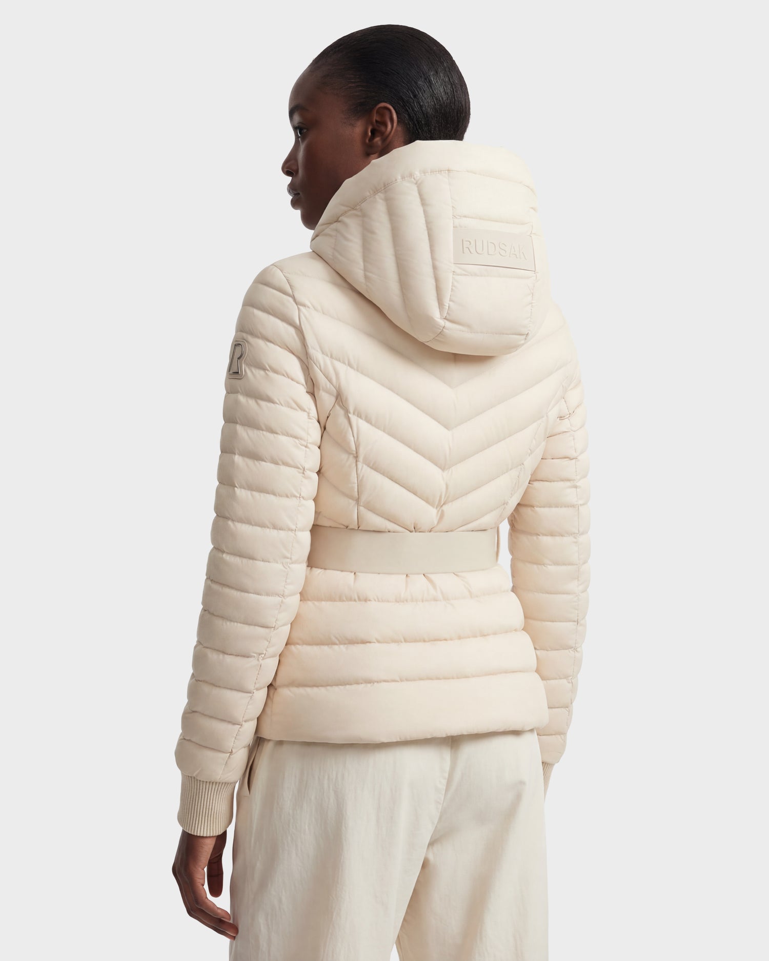 A woman is seen from the back wearing the Rudsak SCARLETT - 8125004 Vanilla light beige, water-resistant, quilted hooded puffer jacket with “RUDSAK” embroidered on the hood and chevron stitching, paired with matching light pants.