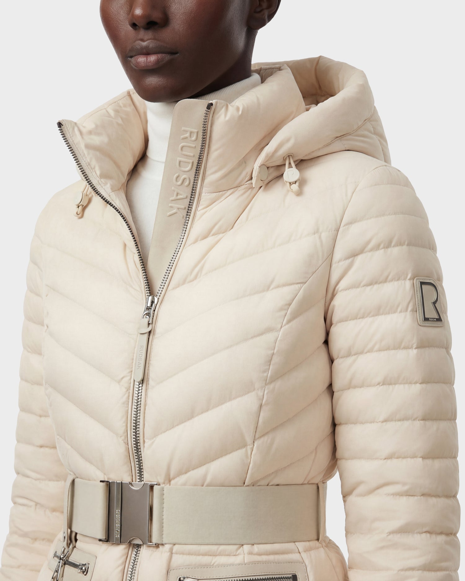 A person wears the Rudsak SCARLETT - 8125004 Vanilla women’s lightweight down jacket in light beige with a high collar, hood, large vegan leather belt, silver zippers, RUDSAK branding, and an "R" patch on the sleeve. Water-resistant design.