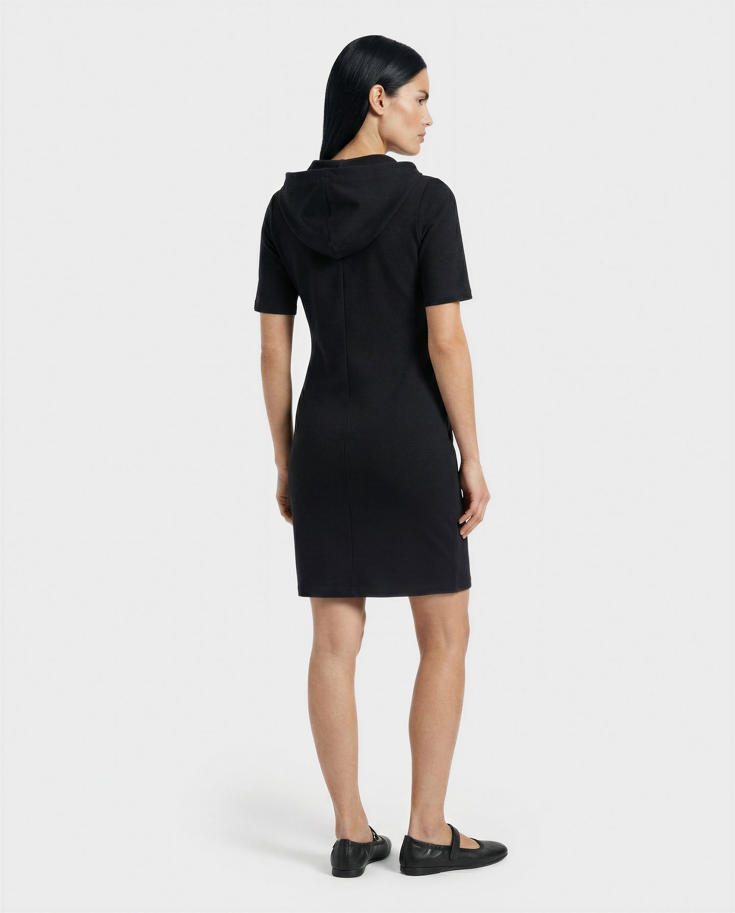 A woman with straight dark hair faces away, wearing the Rudsak GISELA - L524004 Black short-sleeve hooded dress and black shoes against a plain white background.
