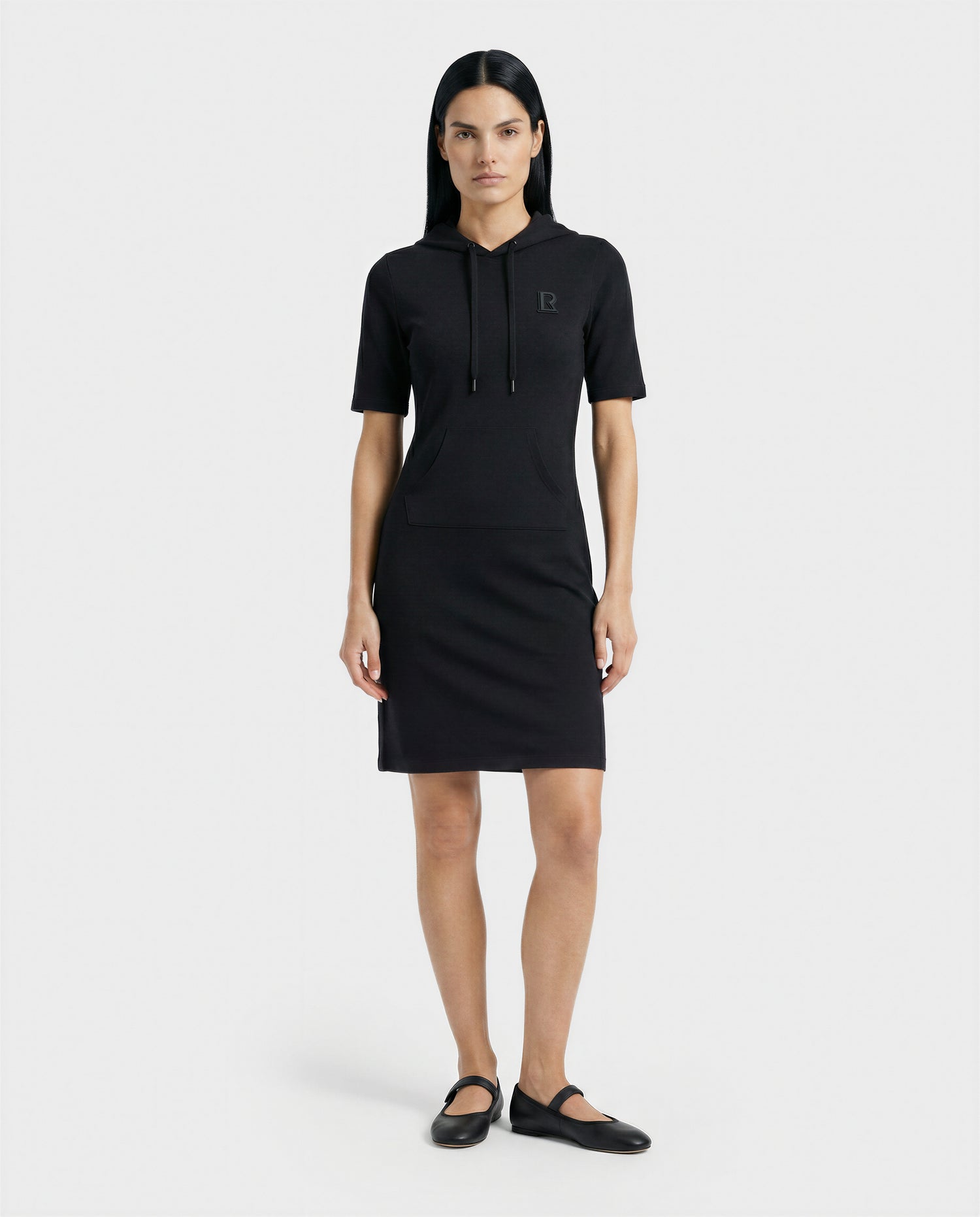 A woman with straight dark hair wears the GISELA - L524004 Black fitted hooded dress by Rudsak, featuring short sleeves, drawstrings, and a front pocket. She stands facing forward in black ballet flats against a white background.