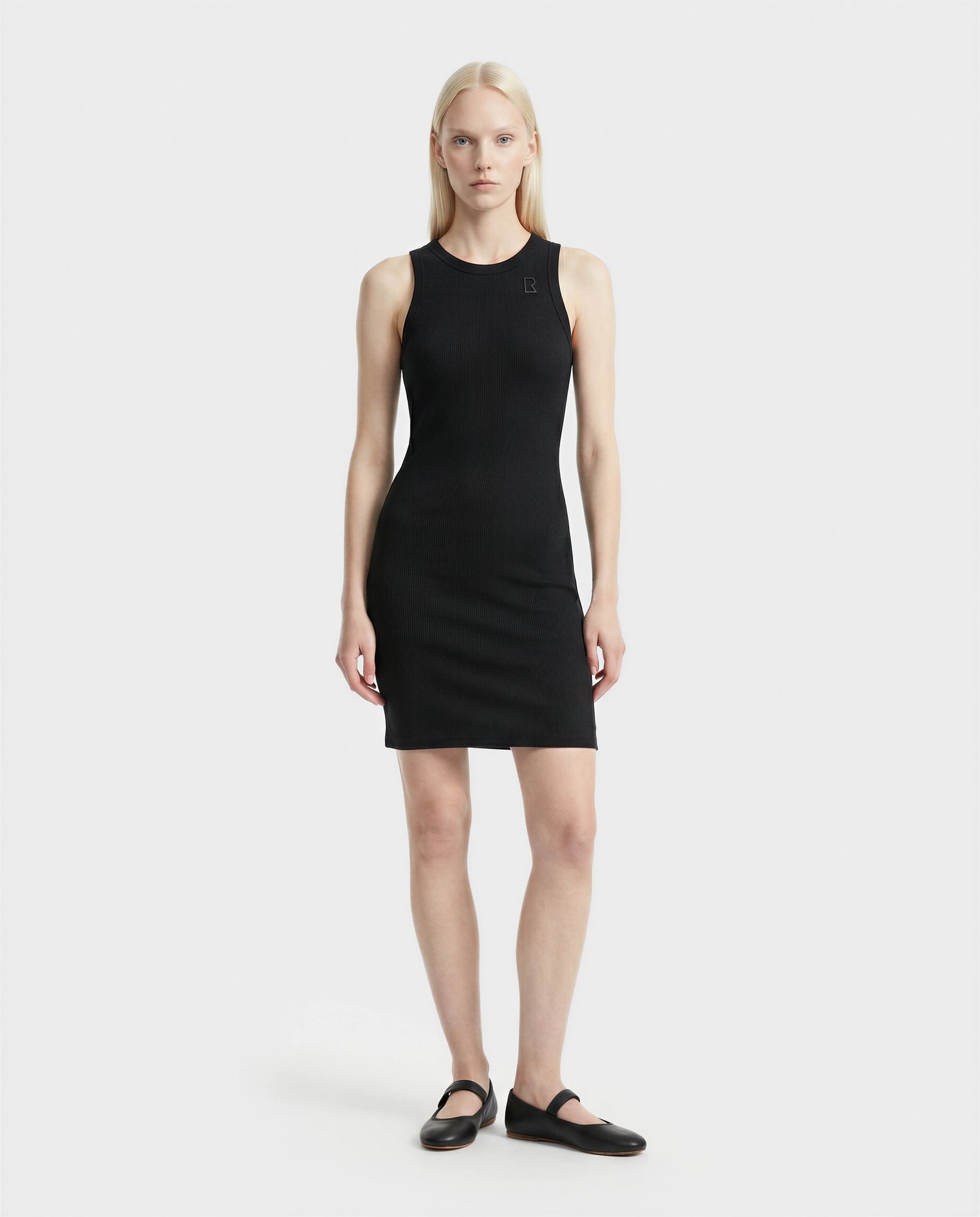 A woman with straight, light blonde hair stands against a white background, wearing the Rudsak ADELLA - L524005 Black sleeveless fitted dress and black flat shoes, facing forward with a neutral expression and arms at her sides.