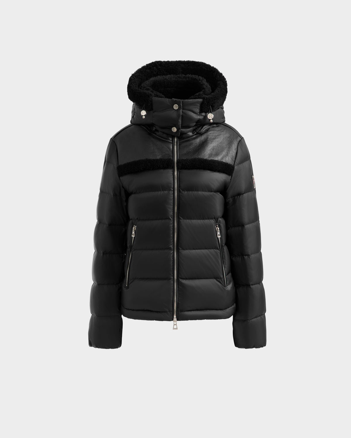 The Rudsak SANDRA - 8125612 Black puffer jacket features lightweight insulation, a hood, front zipper closure, two zippered side pockets, and leather accents on the shoulders and chest, shown on a plain light gray background.