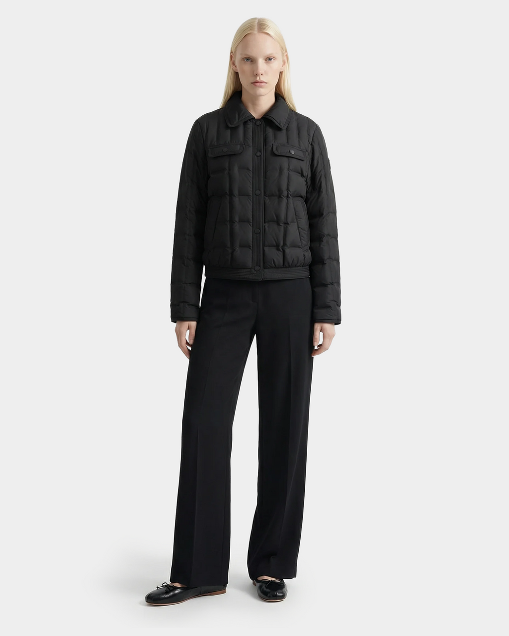 A person with long blonde hair faces forward, wearing the Rudsak SEOUL - 8126027 Black RDS-certified duck down quilted jacket for lightweight warmth, paired with black wide-leg trousers and shoes against a plain light background.