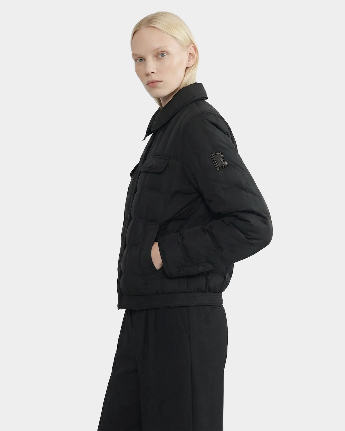 A person with light blonde hair models the Rudsak SEOUL - 8126027 Black quilted down jacket and black pants, standing sideways against a plain light background with a neutral expression.