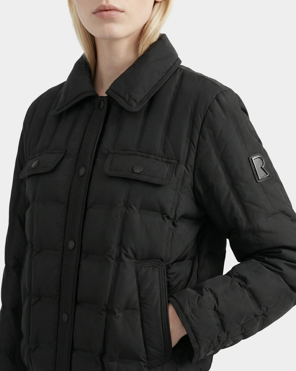 A person with blonde hair, face partially out of frame, wears the Rudsak SEOUL - 8126027 Black quilted down jacket featuring a collar, front flap pockets, R patch on the left sleeve, and lightweight RDS-certified duck down.