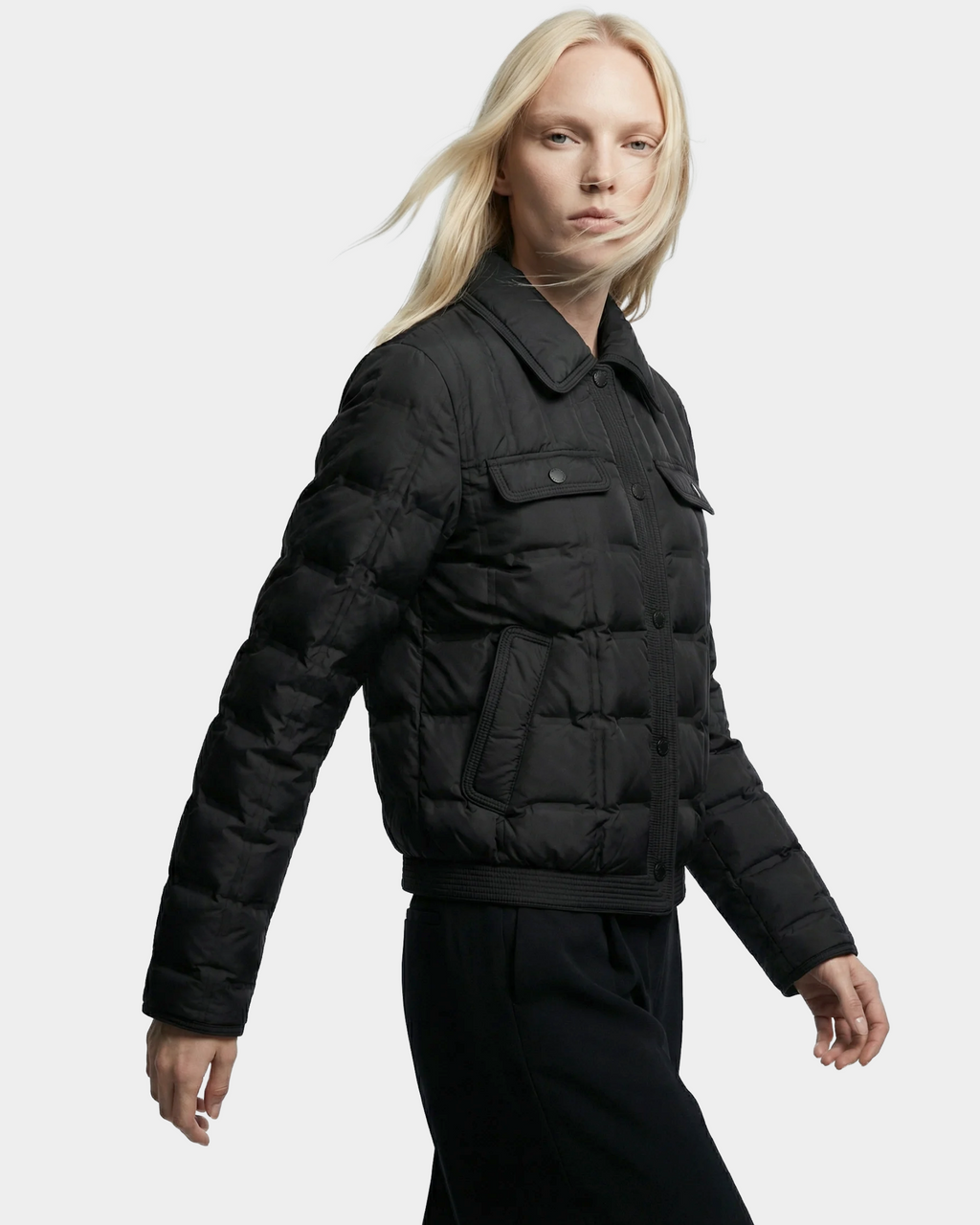 A person with long blonde hair models the Rudsak SEOUL - 8126027 Black quilted down jacket with front pockets and black pants, standing against a light gray background and showcasing its stylish, lightweight warmth.