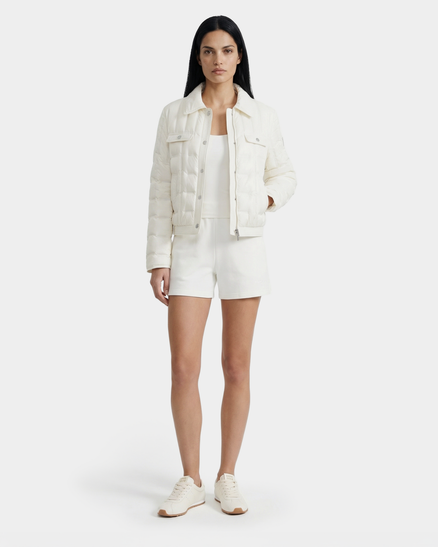A woman with long dark hair models the Rudsak SEOUL - 8126027 Iceland white quilted down shirt jacket, filled with RDS-certified duck down, over a white top and shorts, standing against a light background for lightweight warmth.
